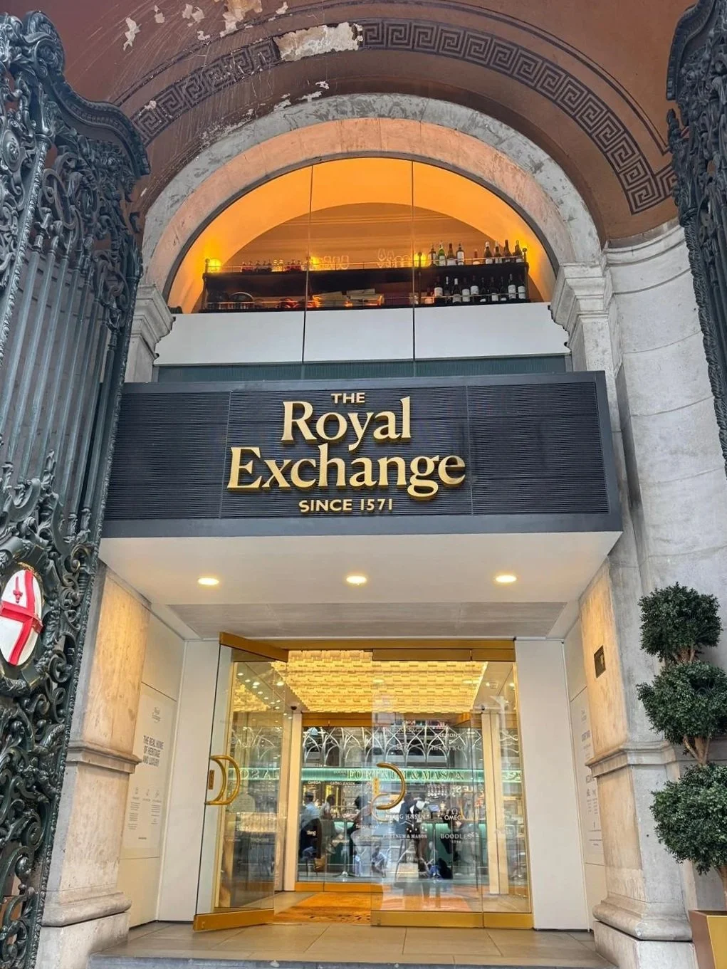 Facade of the Royal Exchange building with a black sign displaying the name, gold lettering, and a glass entrance door with reflections of people and street outside.