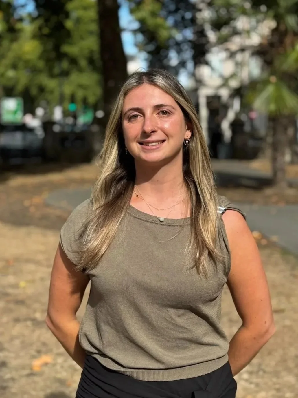 Young woman with long blonde hair standing outside in a park on a sunny day, smiling at the camera, wearing a taupe sleeveless top and black pants.