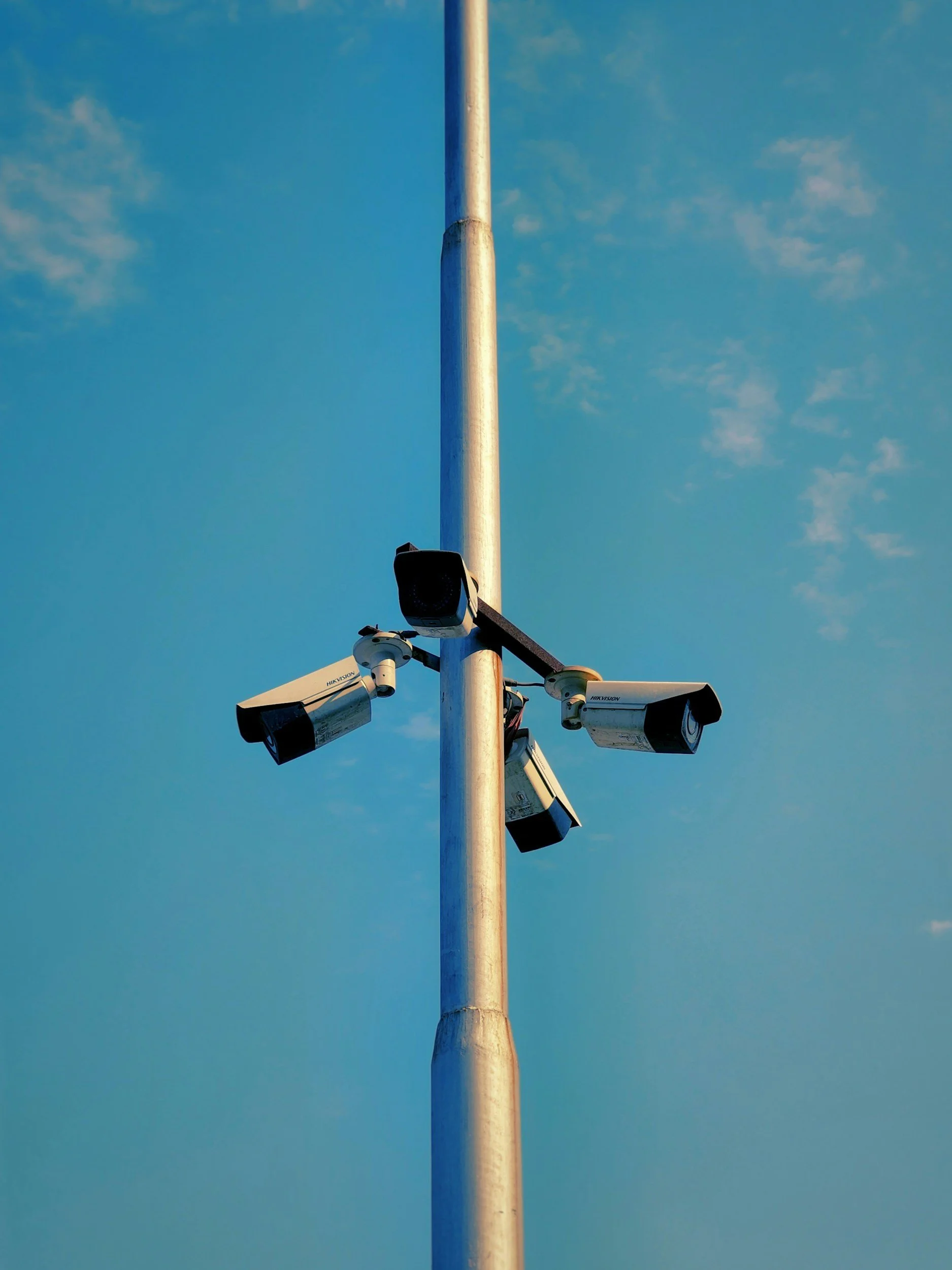 A flagpole with multiple security cameras attached, set against a blue sky with scattered clouds.