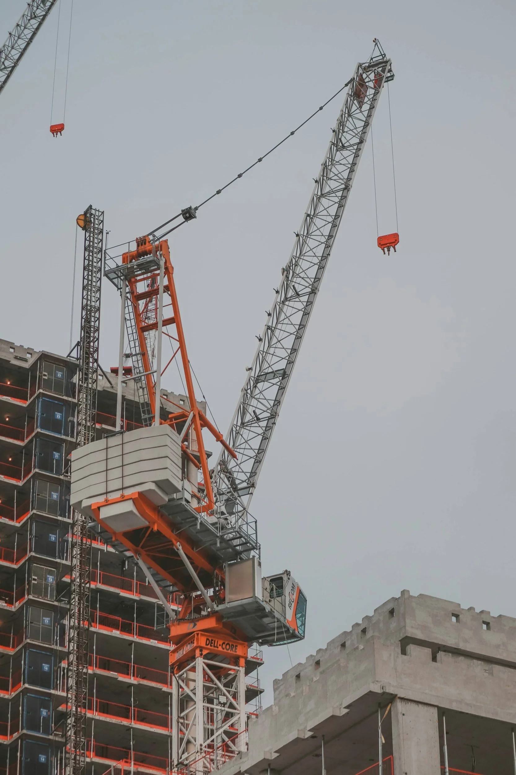 Construction site with two large cranes, one with orange and white colors, lifting materials near a multi-story building under construction, with a nearby gray building against a cloudy sky.