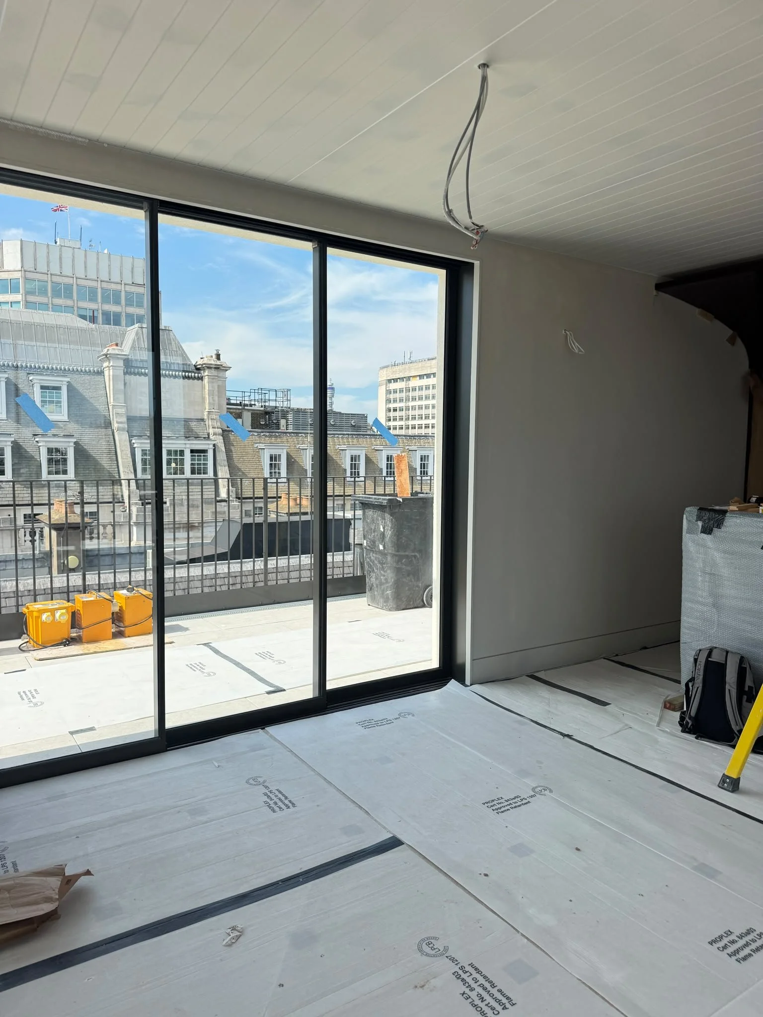 Interior room under construction with large glass sliding door leading to a balcony. The floor is covered with protective sheets, and there are construction materials and tools. Outside, there are buildings with a clear sky.
