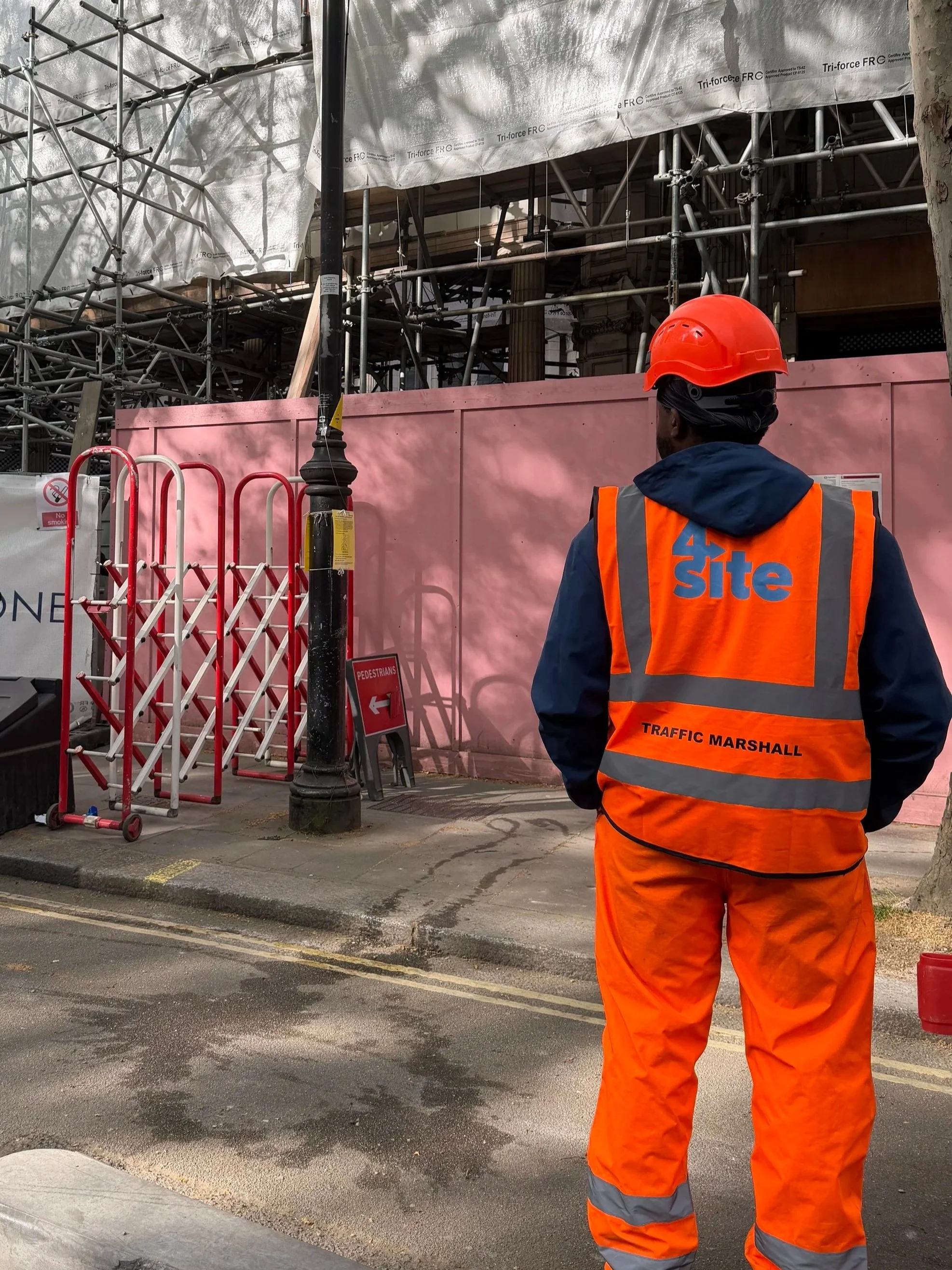 A construction worker wearing an orange safety helmet and high-visibility orange vest standing outside a building under construction, with pink exterior wall panels and scaffolding in the background.