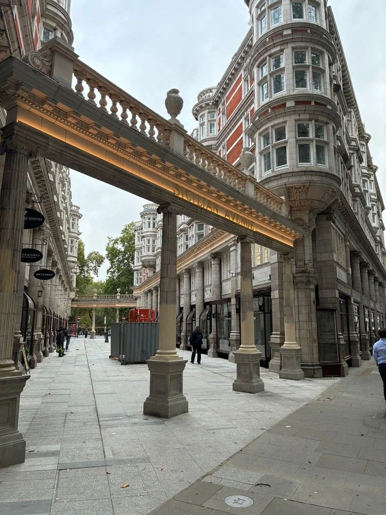 A city street scene with ornate historic buildings, columns supporting an awning with a sign, and a few pedestrians walking on the sidewalk.