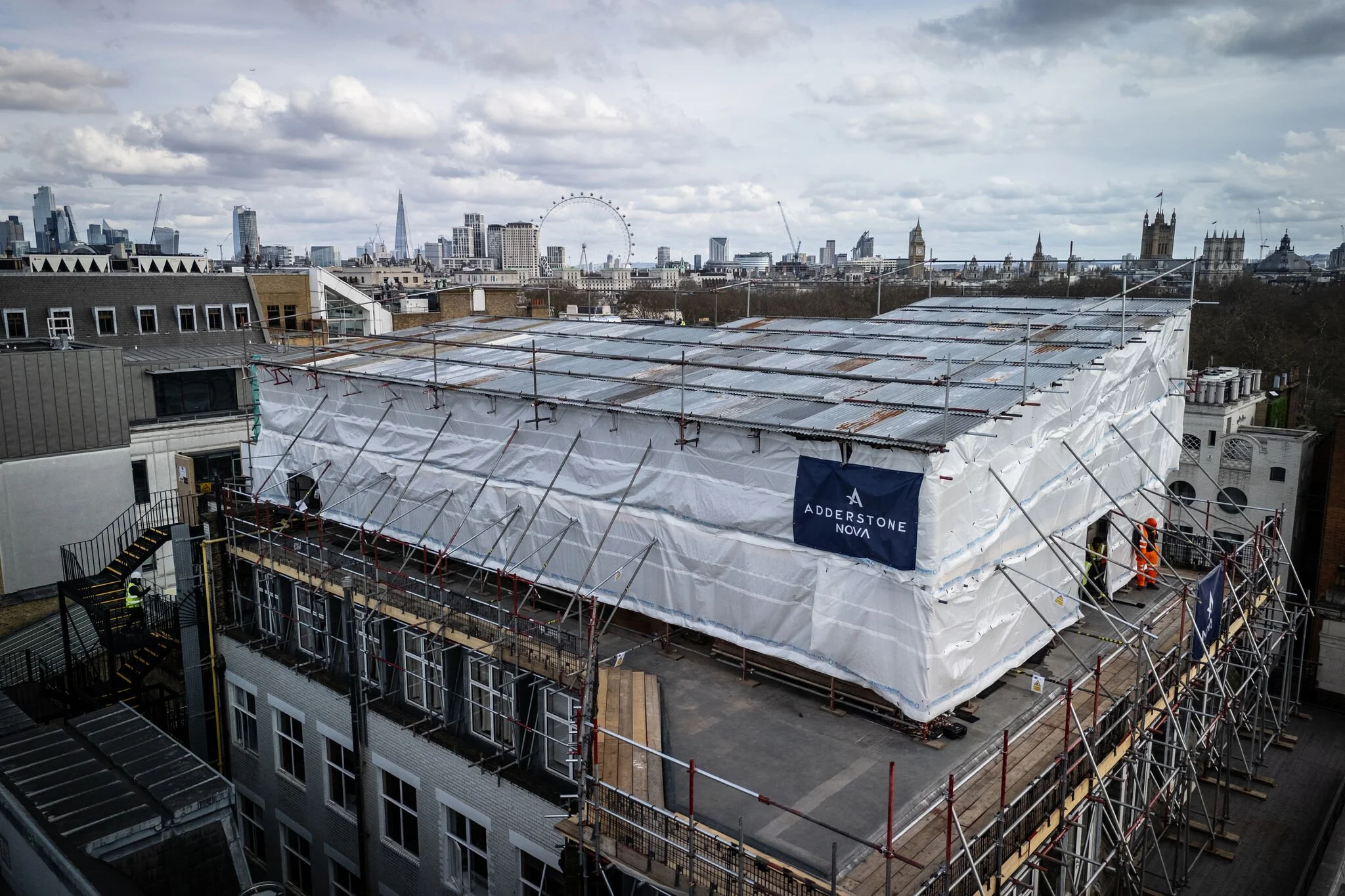 Construction site on a building with scaffolding and white protective covering, city skyline in the background including London landmarks like the London Eye and Big Ben.
