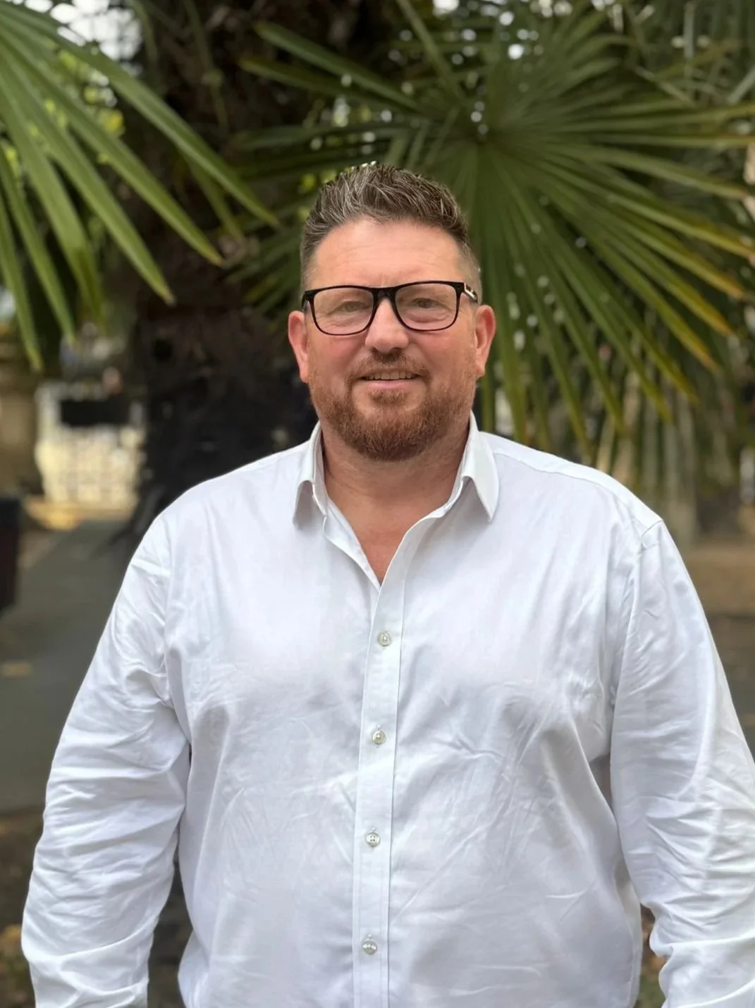 A man with glasses and a beard wearing a white button-up shirt standing outdoors in front of a large palm tree.