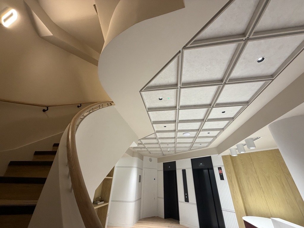 Interior lobby with elevator doors, curved staircase with wooden handrail, white walls, and a ceiling with a gray grid design.