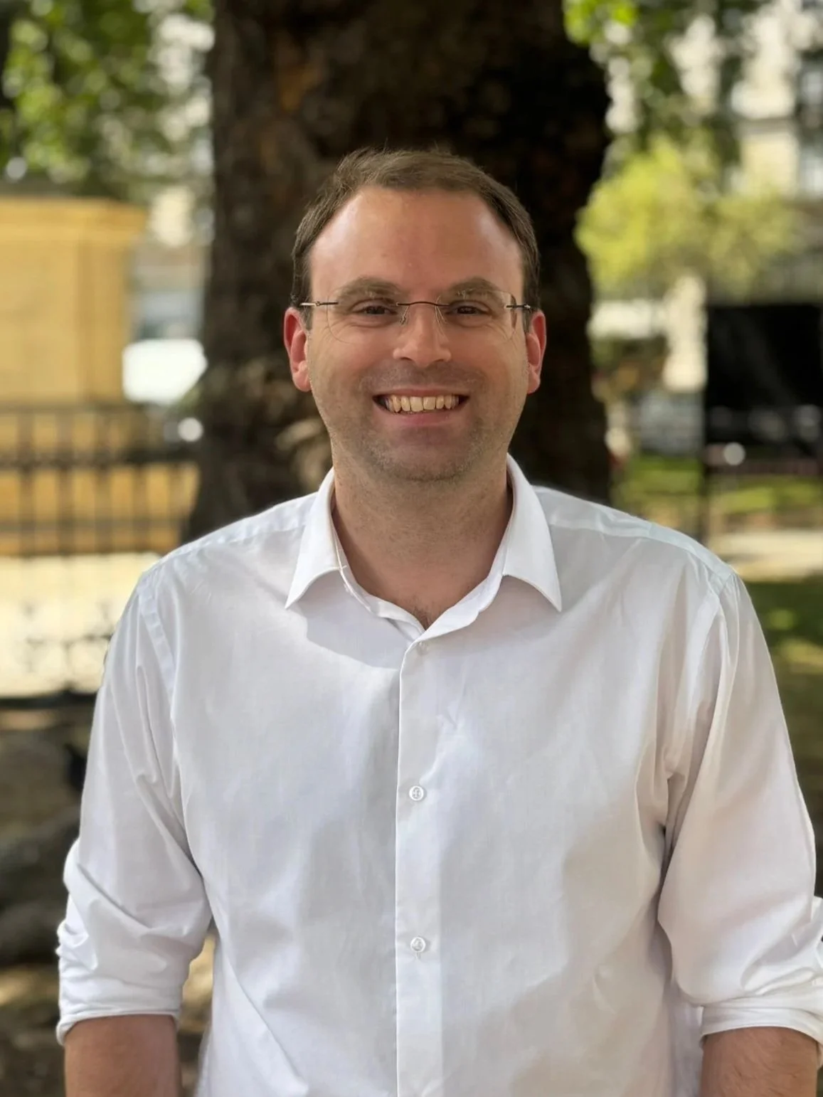 A smiling man in glasses and a white button-up shirt standing outdoors near a large tree.