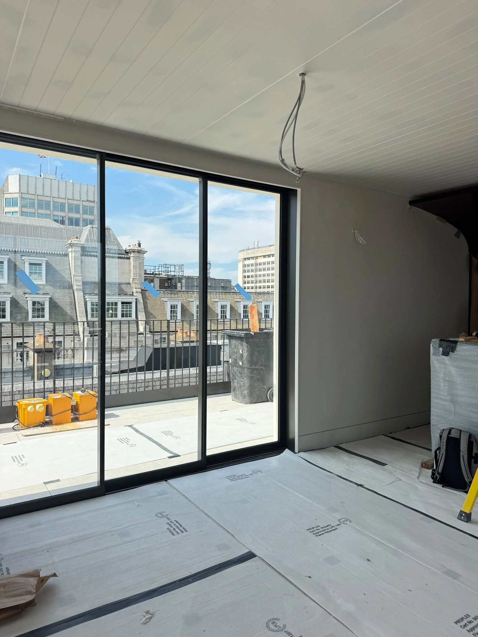 Interior of a high-rise building under construction, featuring large sliding glass doors leading to a balcony with a city skyline view, construction materials, and wiring on the ceiling.
