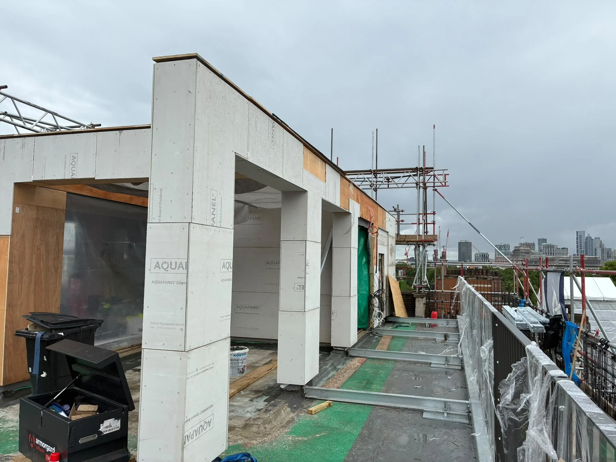Construction site on a rooftop with building framing, scaffolding, and city buildings in the background under cloudy sky.
