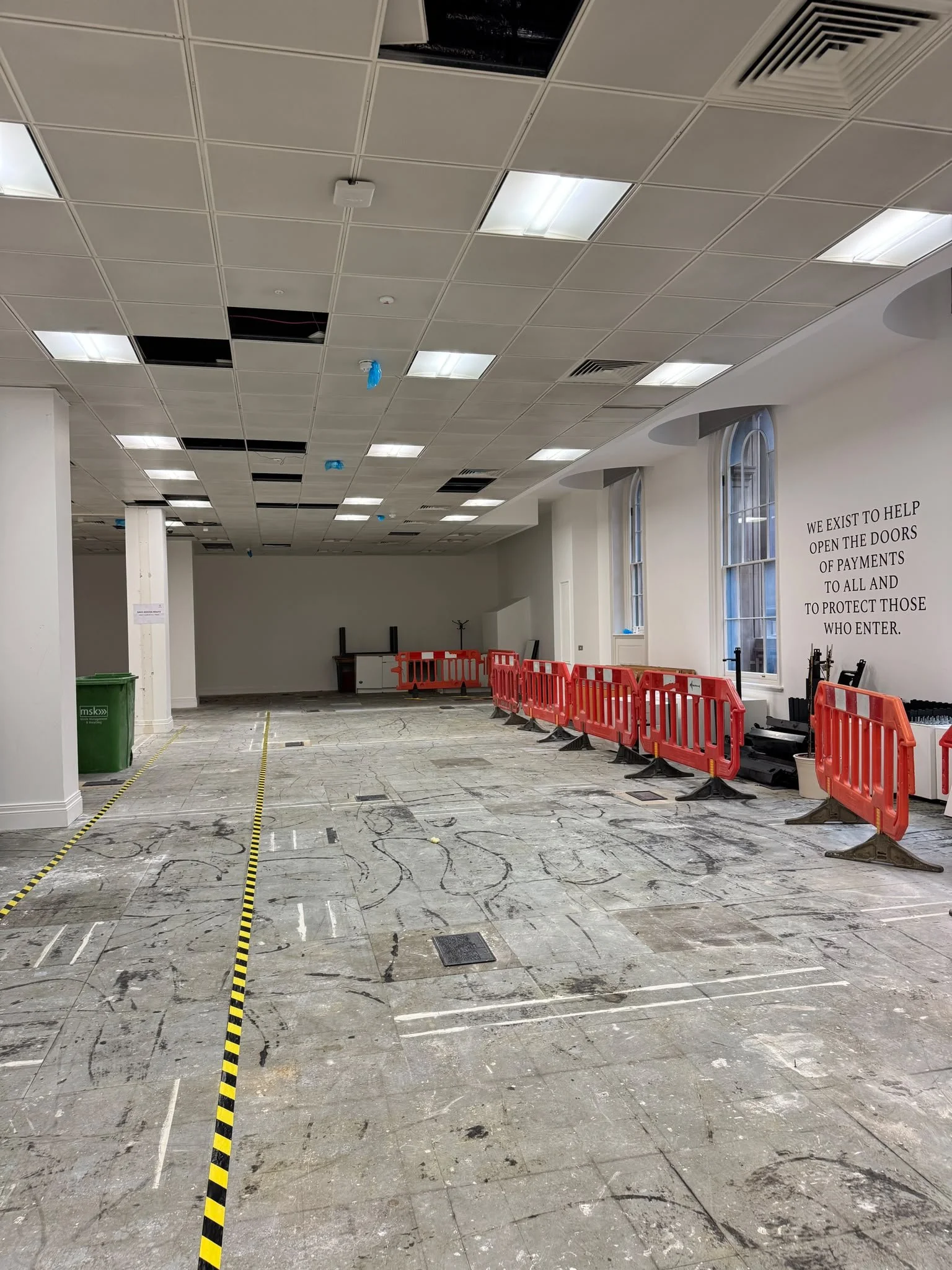 Partially finished interior space with a tiled ceiling, some missing ceiling panels, construction barriers, and a large warehouse-style window, with markings on the floor and a sign on the wall.