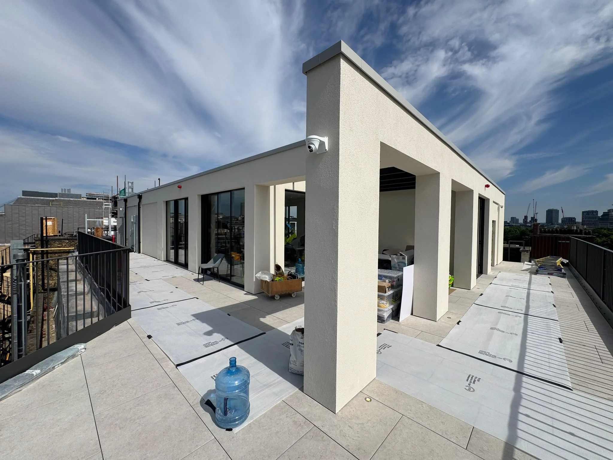 Rooftop terrace under construction with new white building, glass doors, construction materials, water bottles, and city skyline in the background.