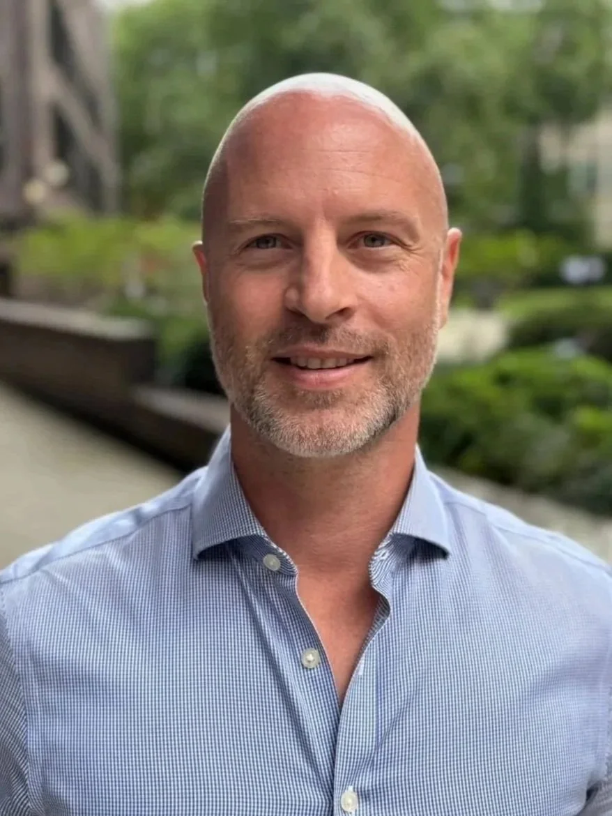 A middle-aged man with a bald head, blue eyes, and a short beard, wearing a light blue dress shirt, smiling outdoors with a blurred background of greenery and buildings.