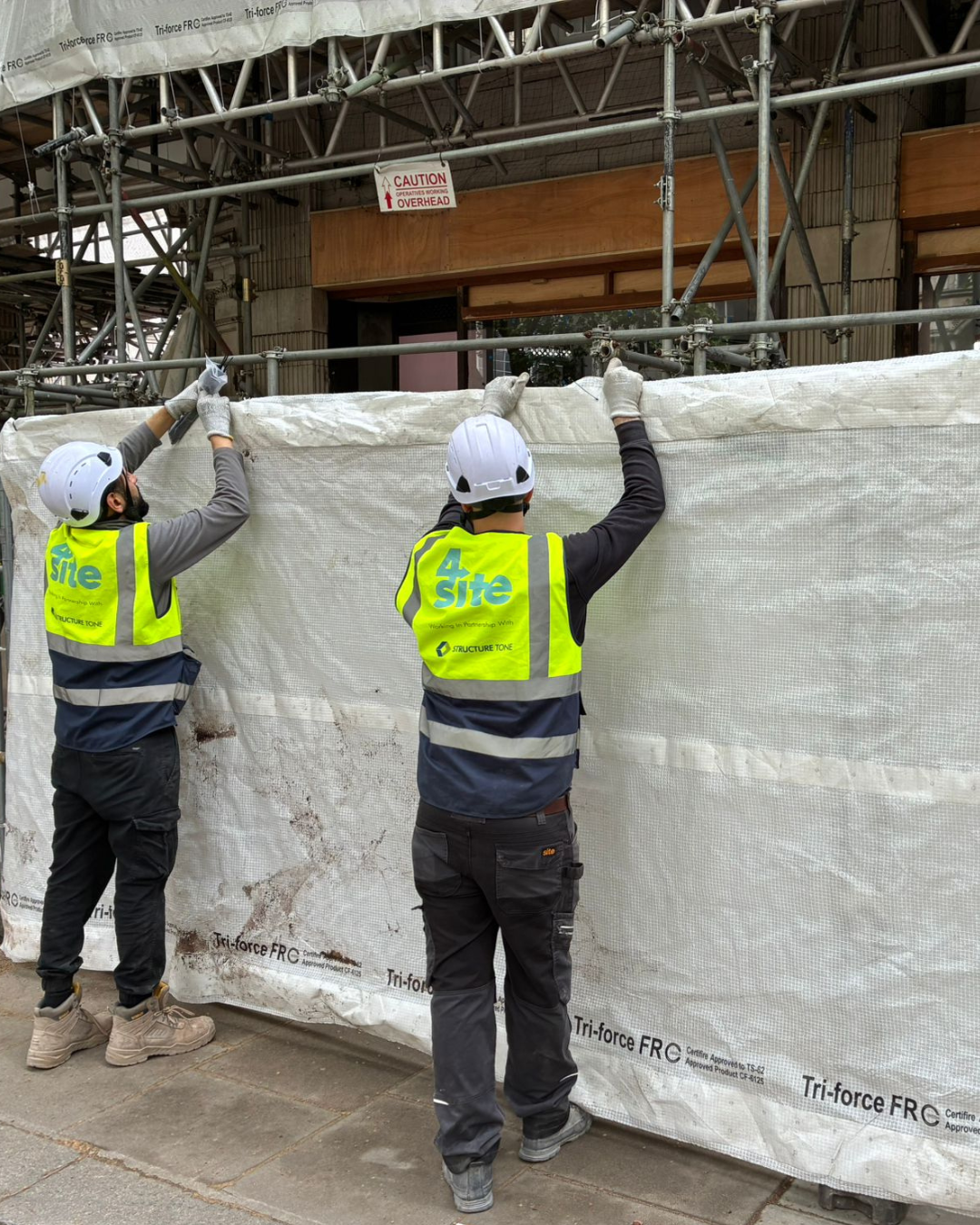 Two construction workers in safety vests and helmets installing or adjusting a white protective barrier on a construction site with scaffolding.