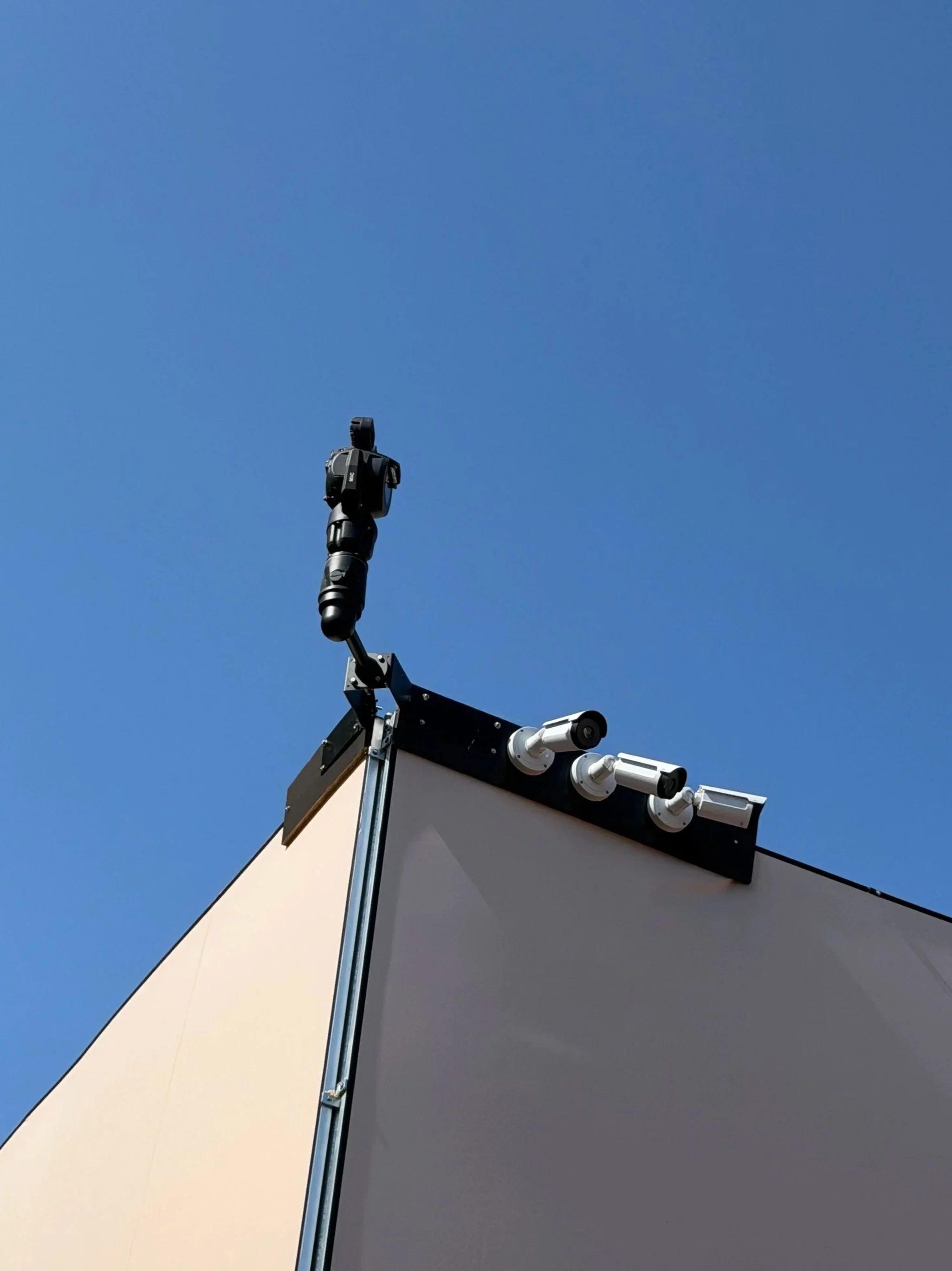 Close-up of a security camera mounted on a corner of a building, with a clear blue sky in the background.