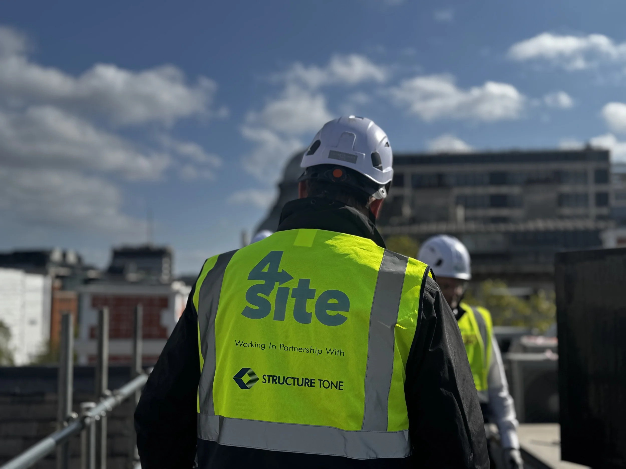 A construction worker wearing a white helmet and a bright yellow safety vest, standing outdoors against a backdrop of a modern building and a blue sky with scattered clouds.