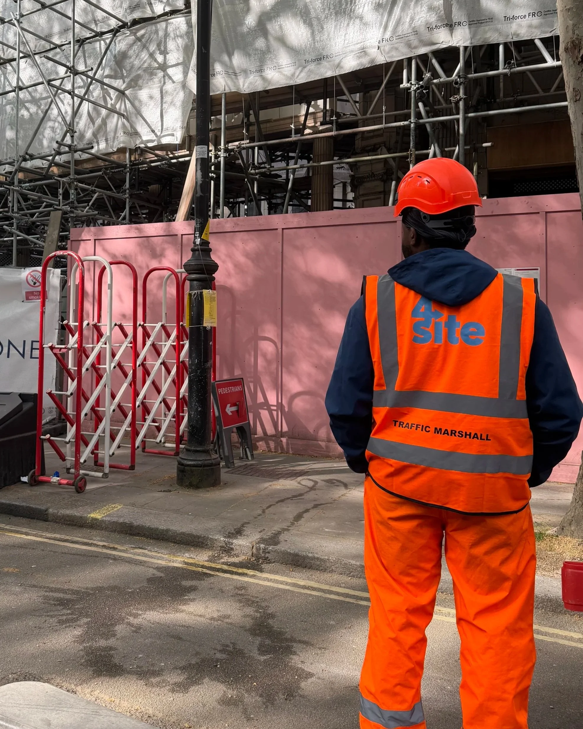 A construction worker wearing a bright orange safety vest and helmet standing on a city street in front of construction scaffolding, a pink barrier, and a no smoking sign.