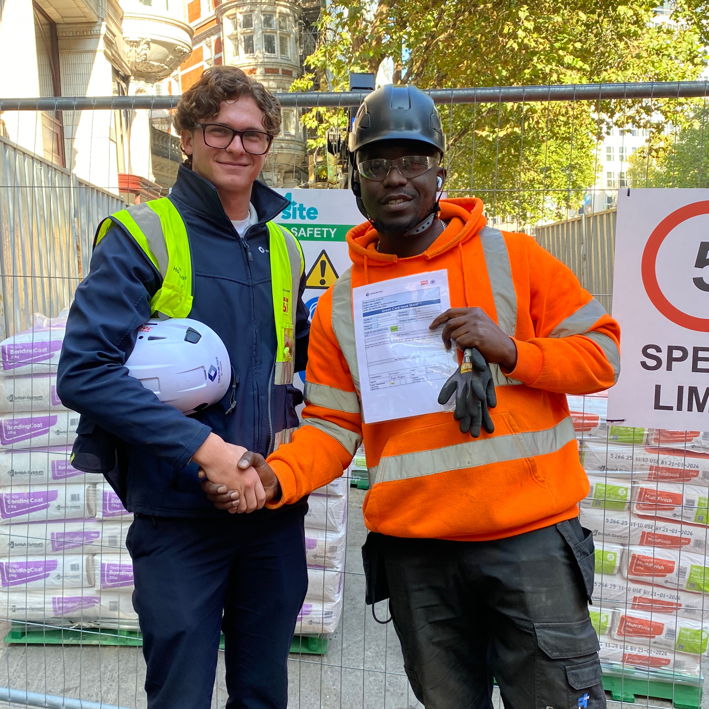 Two men shaking hands at a construction site, one wearing glasses, a navy jacket, and a yellow safety vest, the other wearing a safety helmet, safety glasses, an orange hoodie, and a high-visibility vest, holding a document, with construction materials and safety signs in the background.