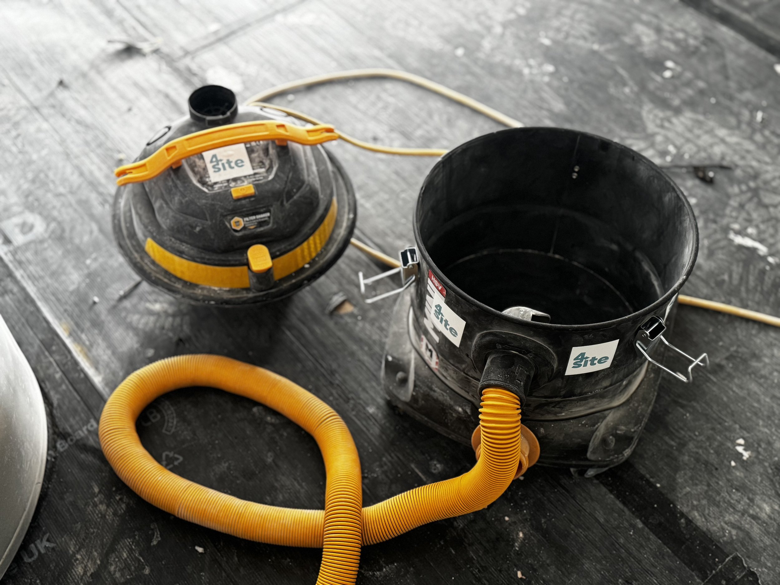 Two black and yellow portable wet/dry vacuums with yellow hoses attached, placed on a dark floor, possibly at a construction or cleaning site.