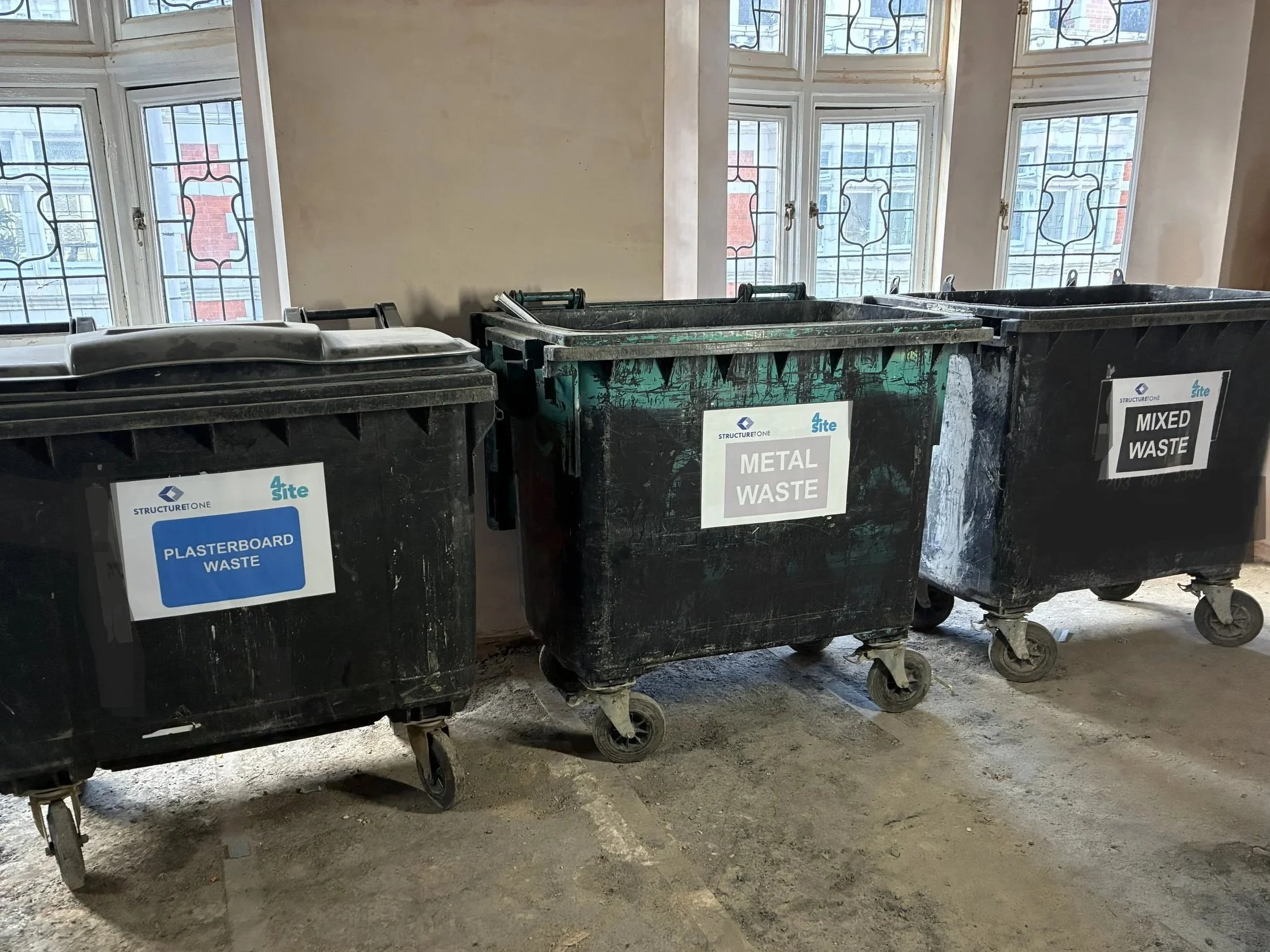 Four large black trash bins labeled for different waste types, lined up inside a room near windows with decorative metalwork, sitting on a concrete or dirt floor.