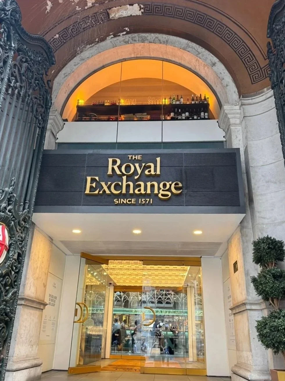 Exterior view of The Royal Exchange building with a sign reading 'The Royal Exchange Since 1571' in London, featuring ornate ironwork gates and a glass entrance, with a restaurant or bar visible inside.