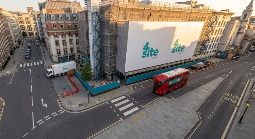Urban construction site with a large billboard on a building under renovation, a red double-decker bus on the road, and surrounding city streets and buildings.