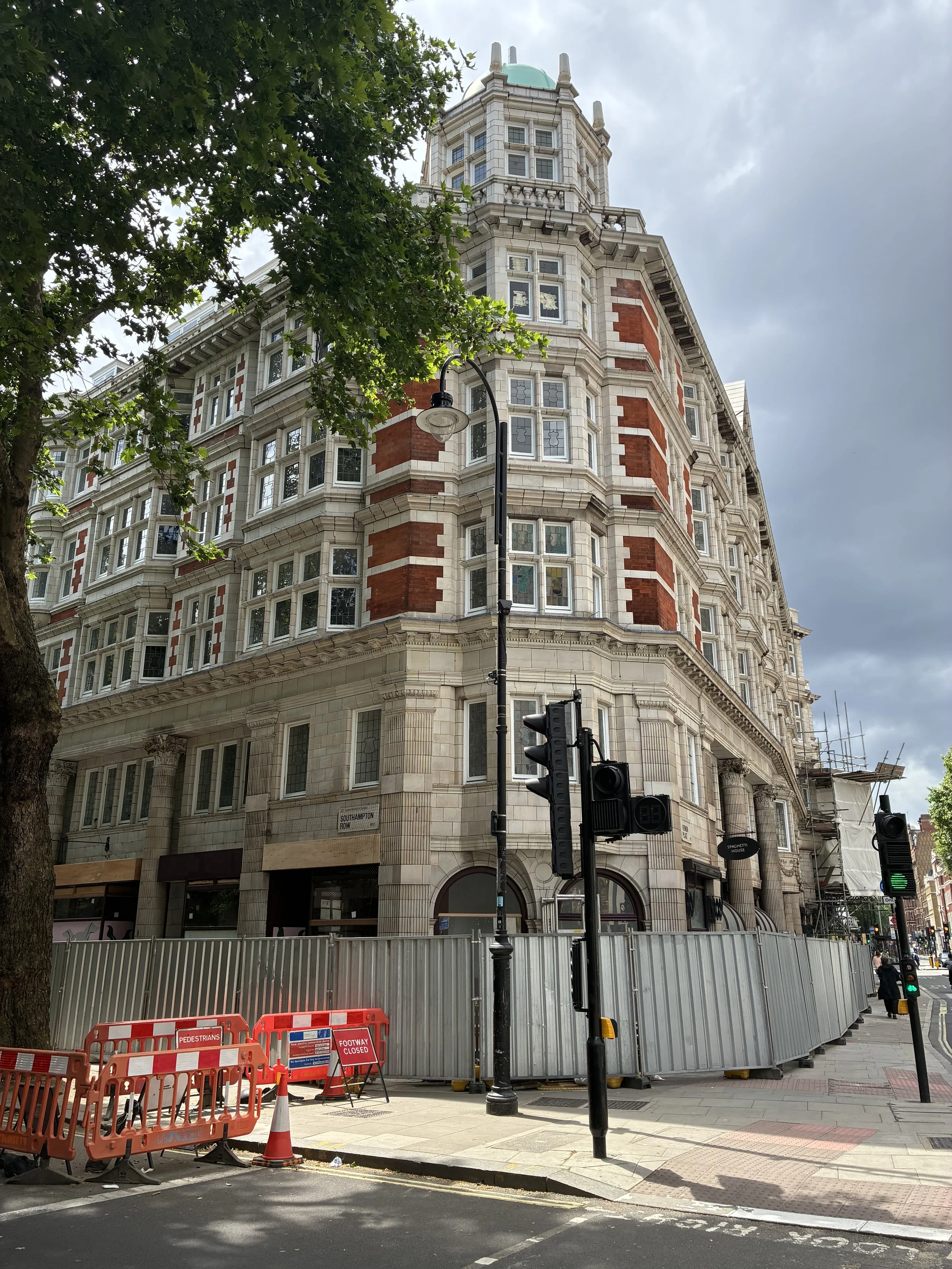 Street view of a large historic building with red brick and white stone details, with scaffolding on part of the building. There are traffic lights, construction barriers, and a tree in the foreground.
