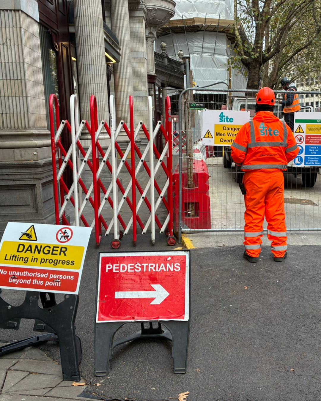 Construction workers in orange safety gear are working on a building site with various warning signs, fencing, and equipment, indicating ongoing construction or renovation, with a partially covered building and trees in the background.