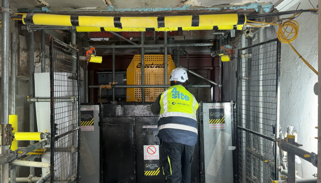 Construction worker inspecting a large piece of heavy equipment inside a building under construction, surrounded by scaffolding and safety barriers.
