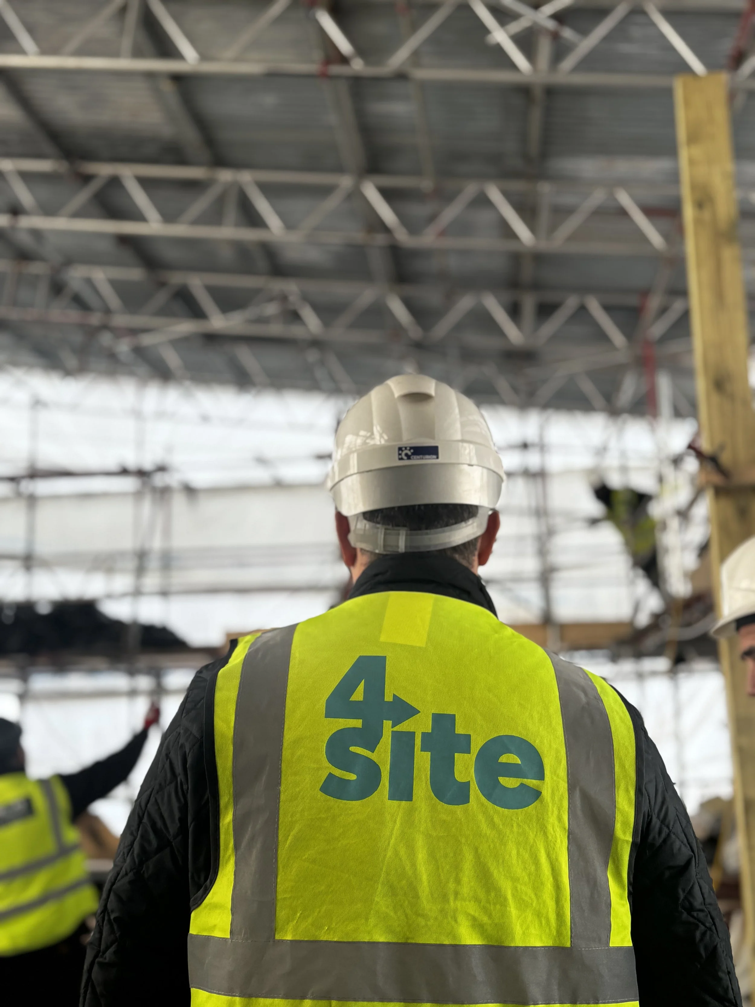 Construction worker wearing a yellow safety vest with the branding of 4Site Logistics on it, standing inside a building under construction with a steel framework ceiling.