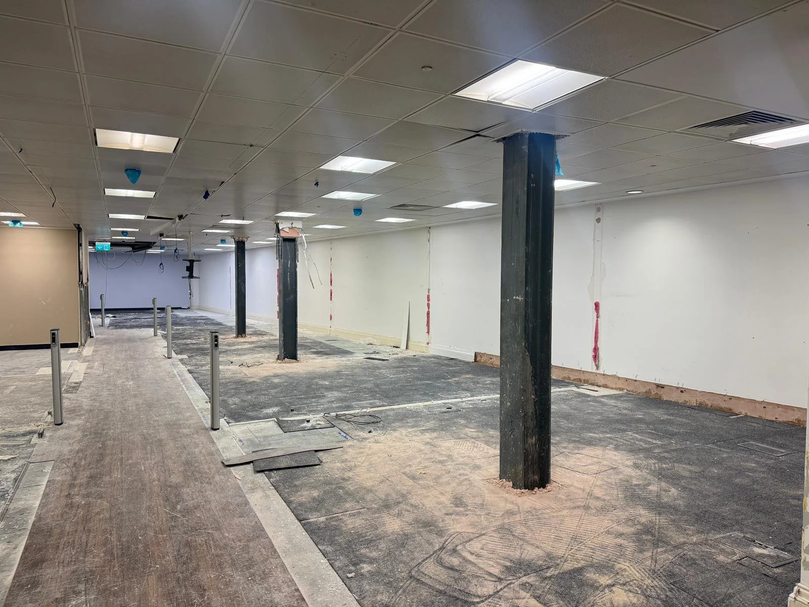 Interior of a building under renovation with exposed support columns, unfinished flooring, and ceiling panels, partially removed wall areas, and construction debris.
