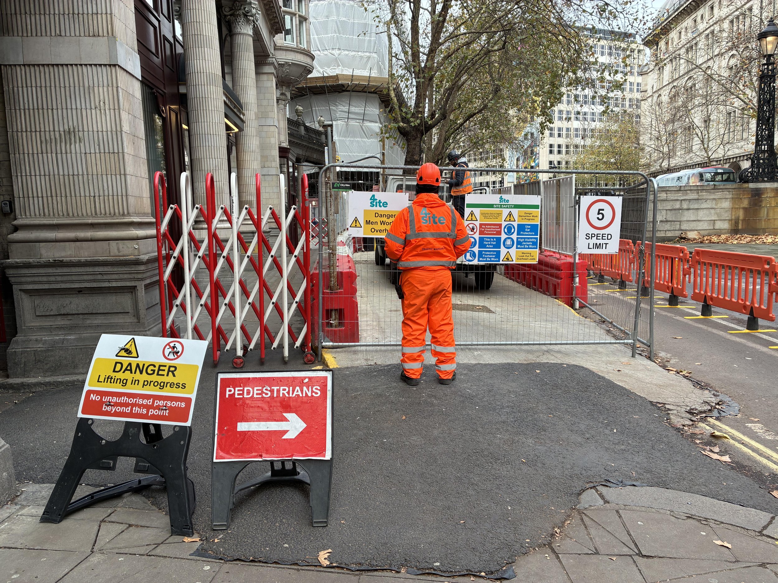 Construction site with workers in orange safety gear, metal fencing, and warning signs including 'Danger Men Working Overhead' and 'Speed Limit 5'. City buildings in the background.