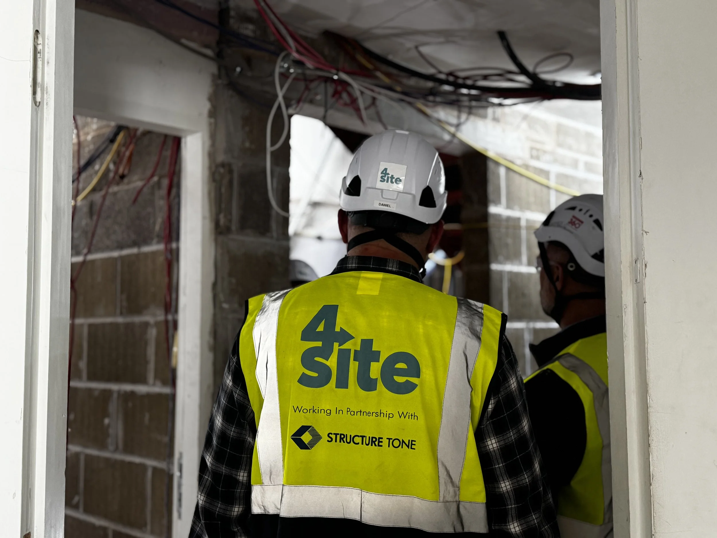 Construction workers wearing yellow safety vests and white helmets inside a partially constructed building with exposed wiring and framing.