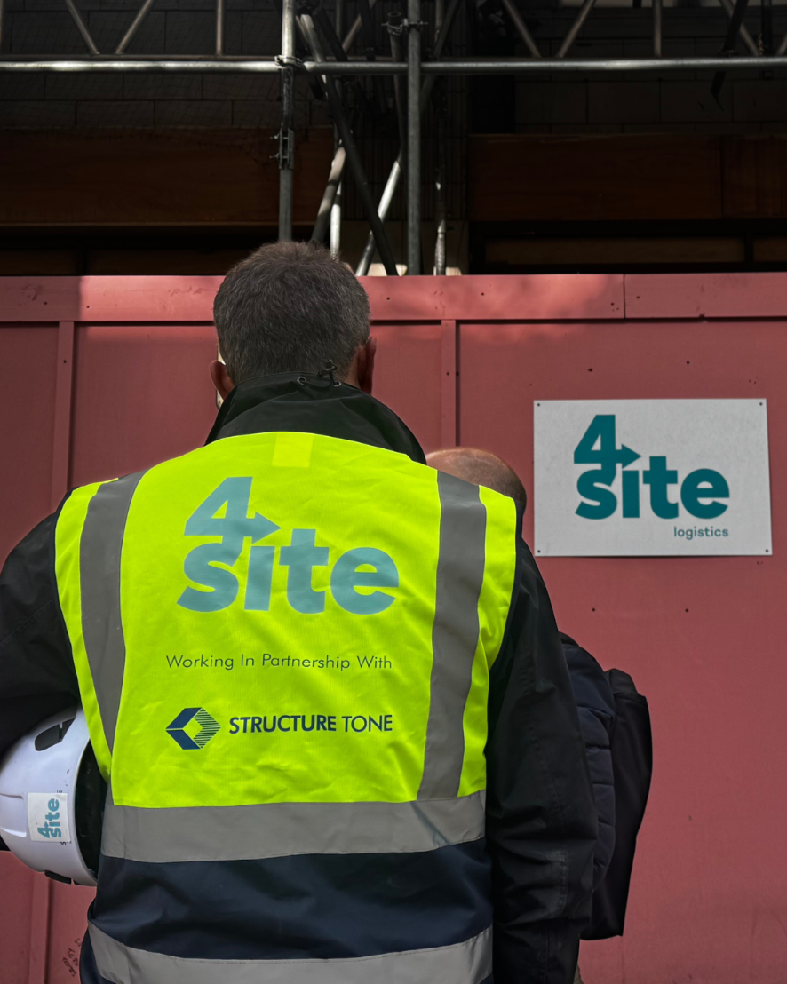 A person wearing a bright yellow safety vest with the logo and name of '4 Site' on the back, standing in front of a pink construction barrier, holding a white helmet, with a sign showing '4 Site logistics' attached to the barrier.