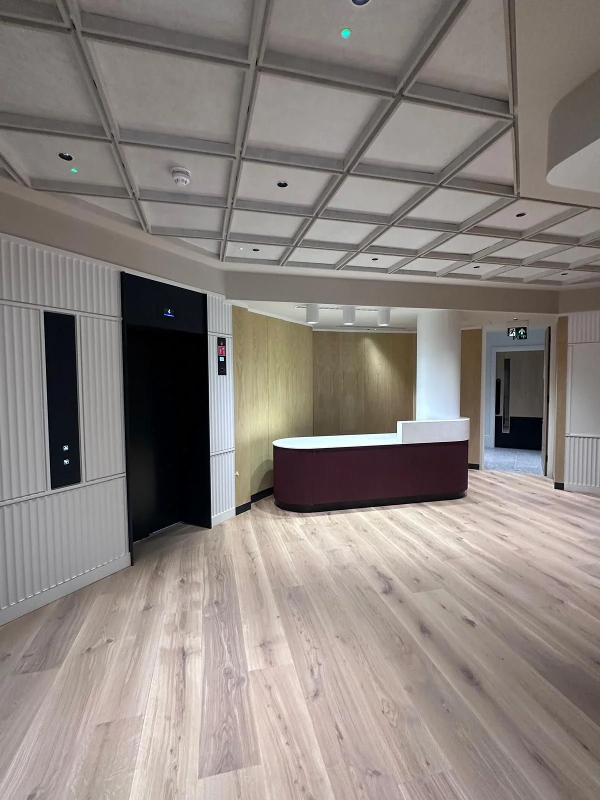 Modern hotel lobby with wood flooring, an elevator with black doors, a curved reception desk with a maroon front, and a hallway with an emergency exit sign.