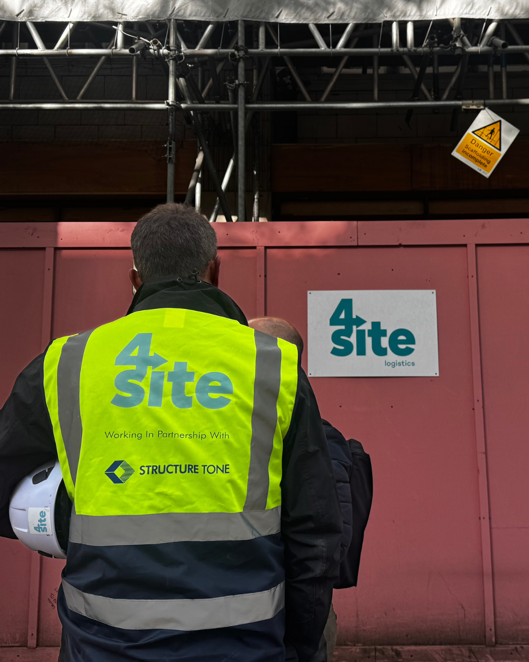 Construction site with a man in a neon yellow safety vest (with '4 Site' and 'STRUCTURE TONE' written on the back) holding a white hard hat. The site is enclosed by a pink barrier with a sign that reads '4 Site logistics.' In the background, there is scaffolding and a warning sign indicating ongoing, incomplete scaffolding work.