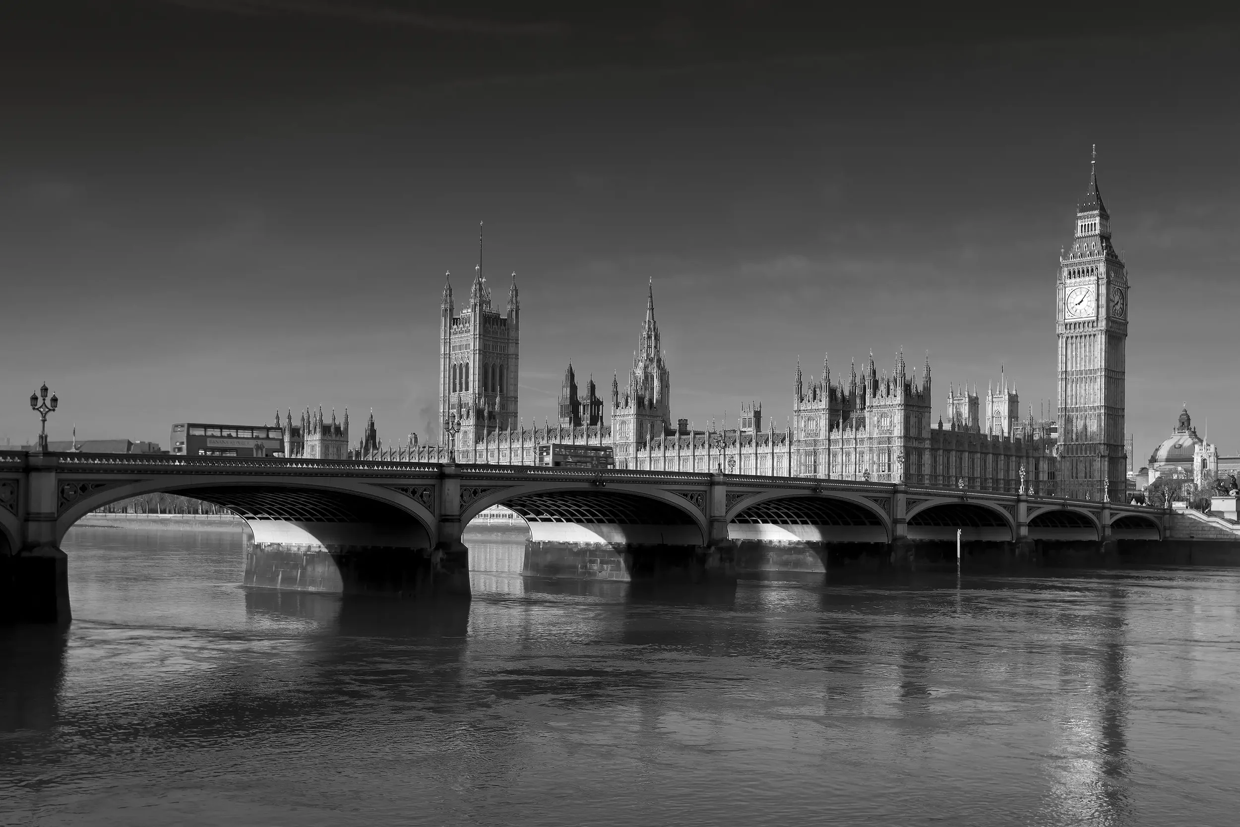 Black-and-white photo of the Houses of Parliament and Big Ben in London, with a bridge crossing the River Thames in the foreground.