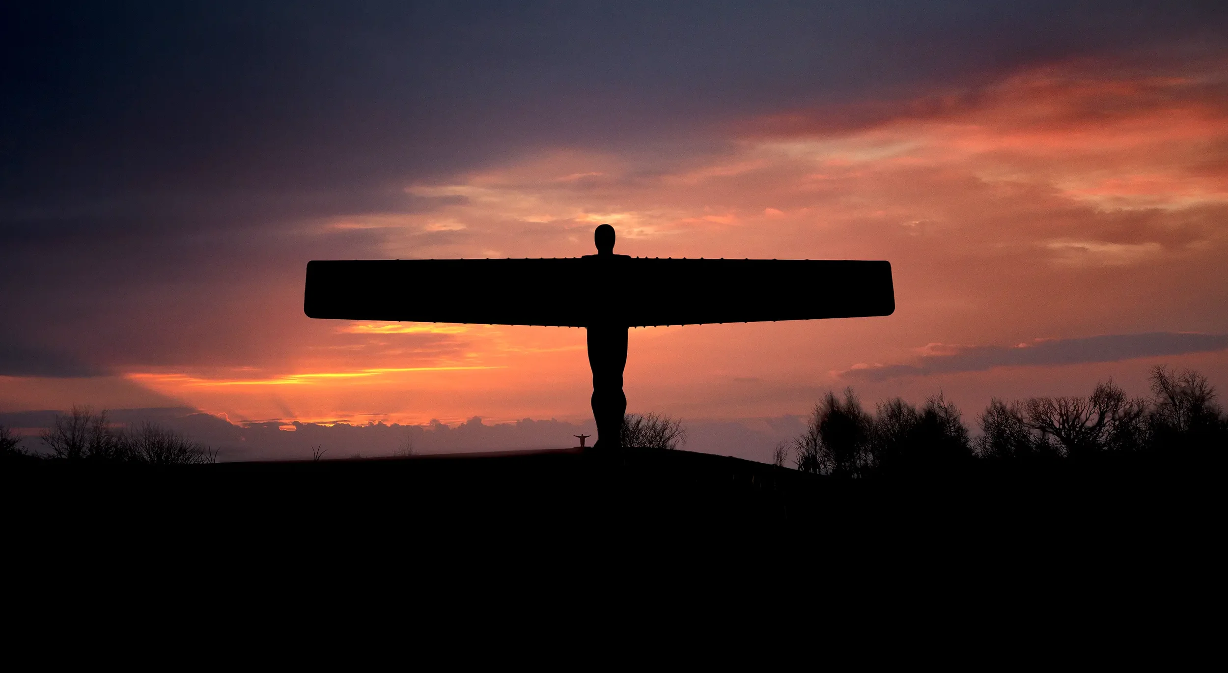 Silhouette of a large cross with a figure on top, set against a colorful sunset sky with clouds and trees in the foreground.