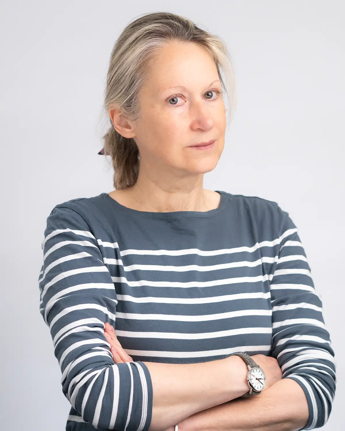 A woman with light-colored hair and fair skin is standing with arms crossed, wearing a gray and white striped long-sleeve shirt and a wristwatch, looking directly at the camera with a neutral expression against a plain light background.