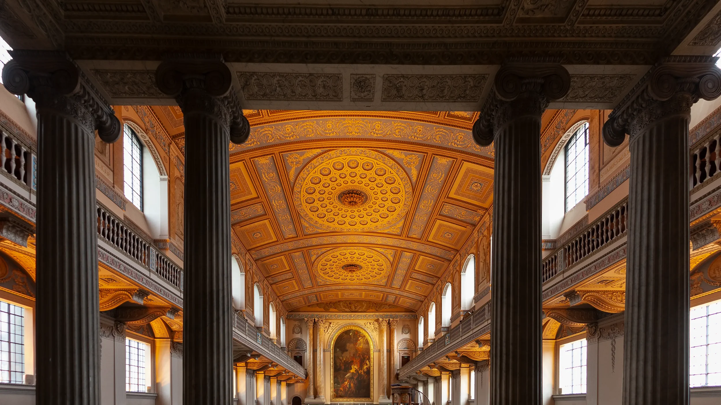 Interior of a grand historic building with tall black columns, ornate gold ceiling, and large arched windows.