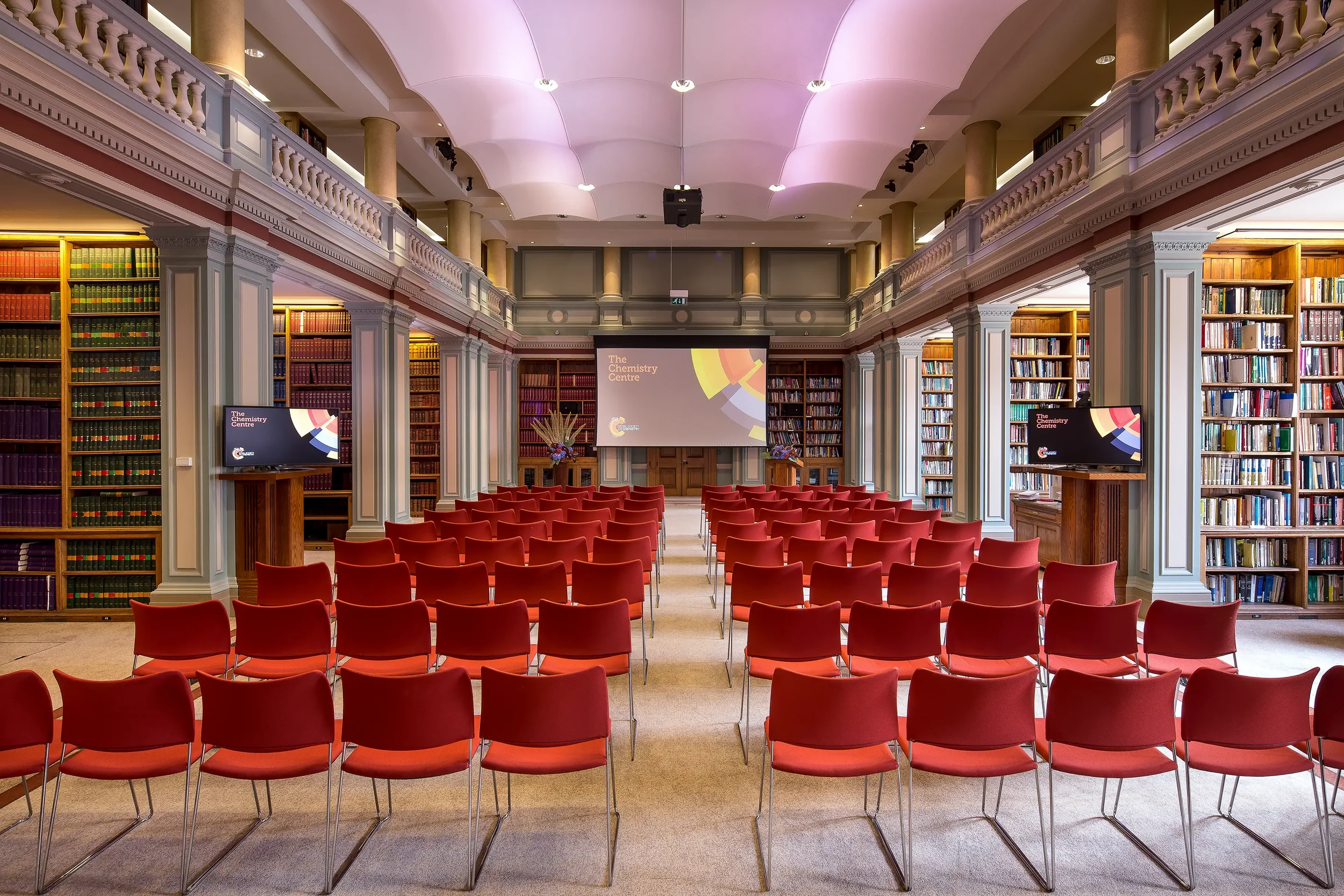 A spacious library conference room with red chairs arranged in rows facing a projector screen that says 'The Chemistry Centre' and a stage with flowers. The room has high ceilings, granite columns, and bookshelves along the walls.