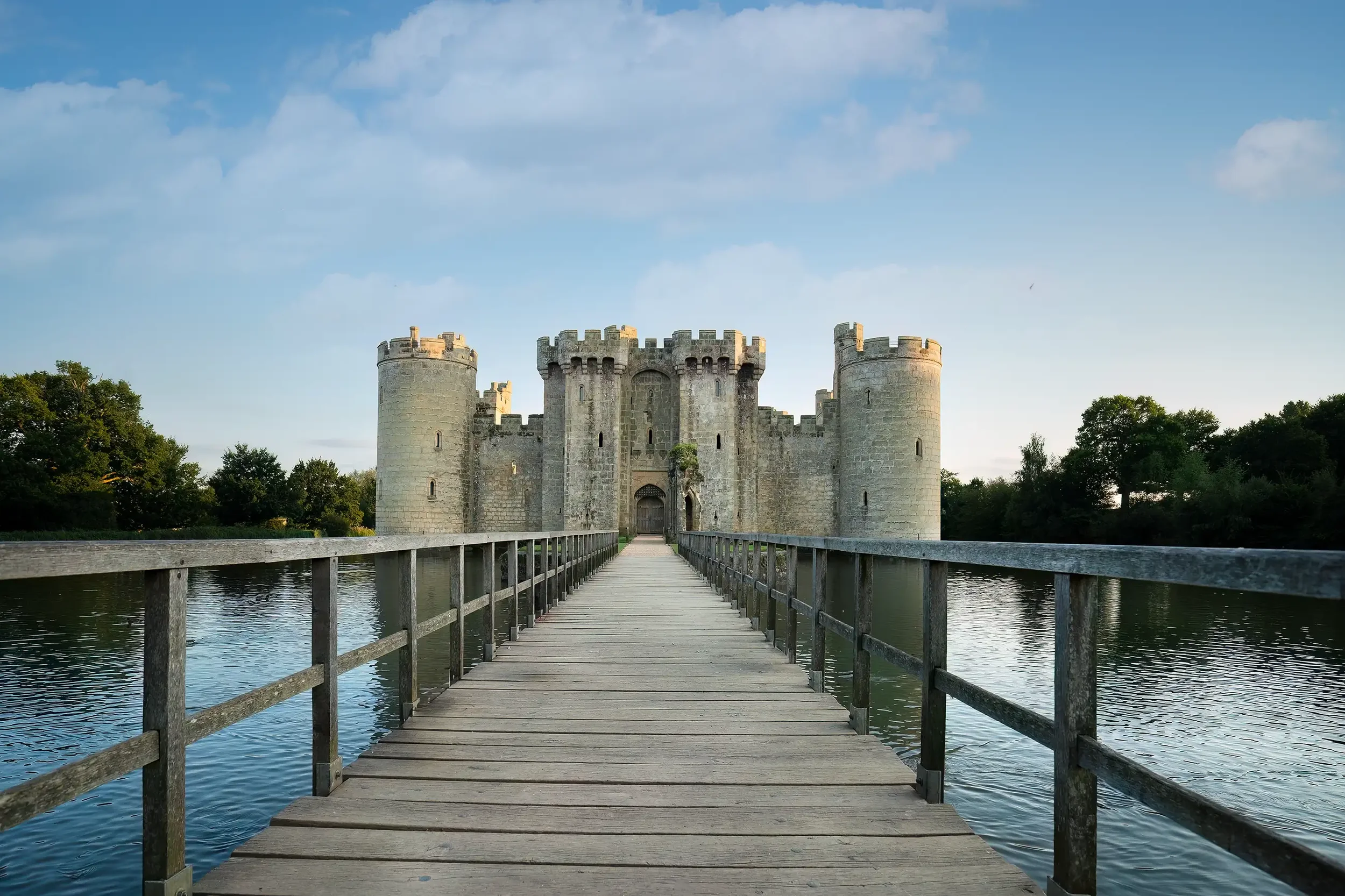 A wooden bridge leading to a medieval stone castle surrounded by water and green trees under a blue sky with clouds.