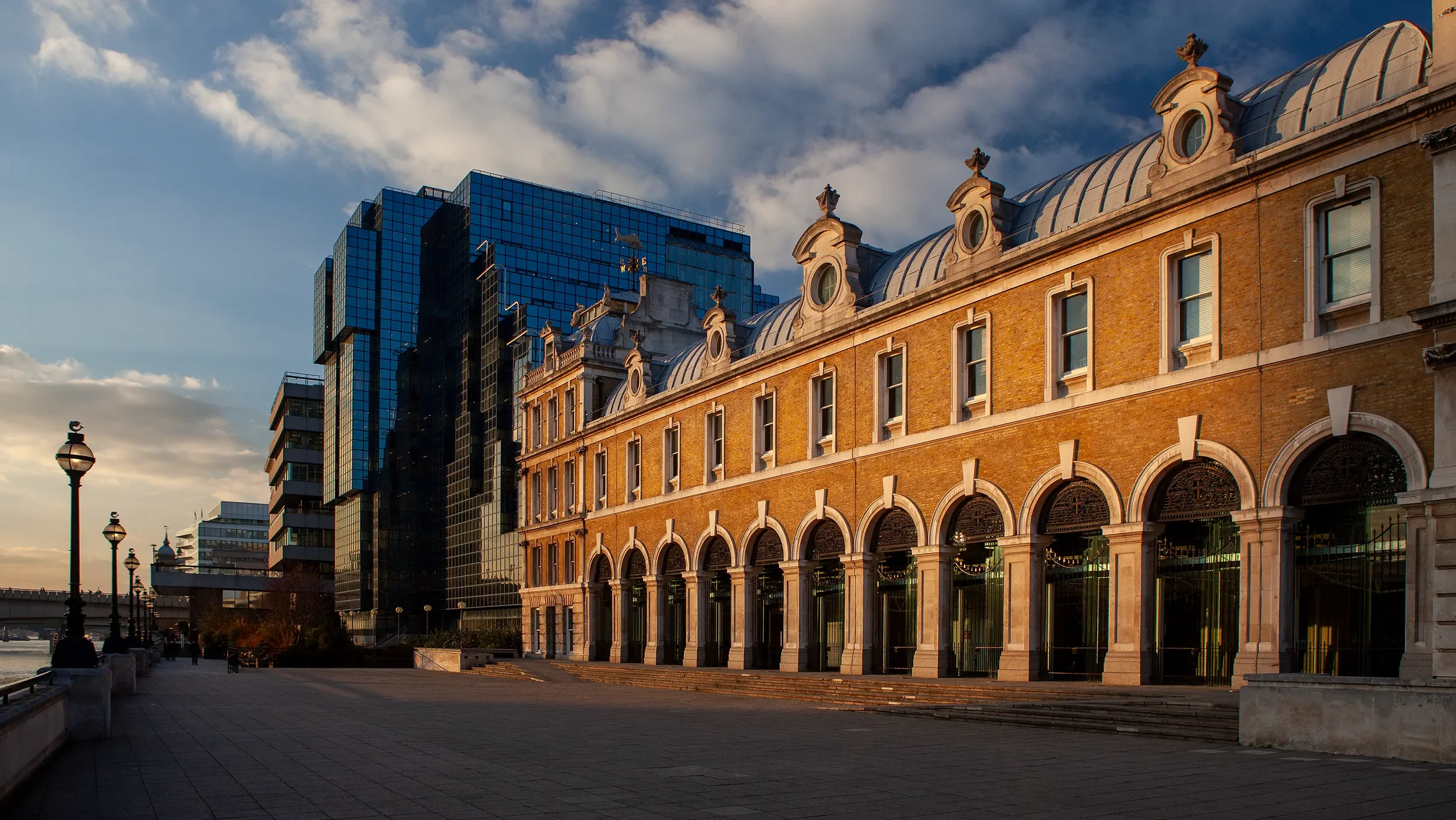 Historical building with arched windows and decorative rooftop elements in front of modern glass skyscrapers, set against a partly cloudy sky in late afternoon.