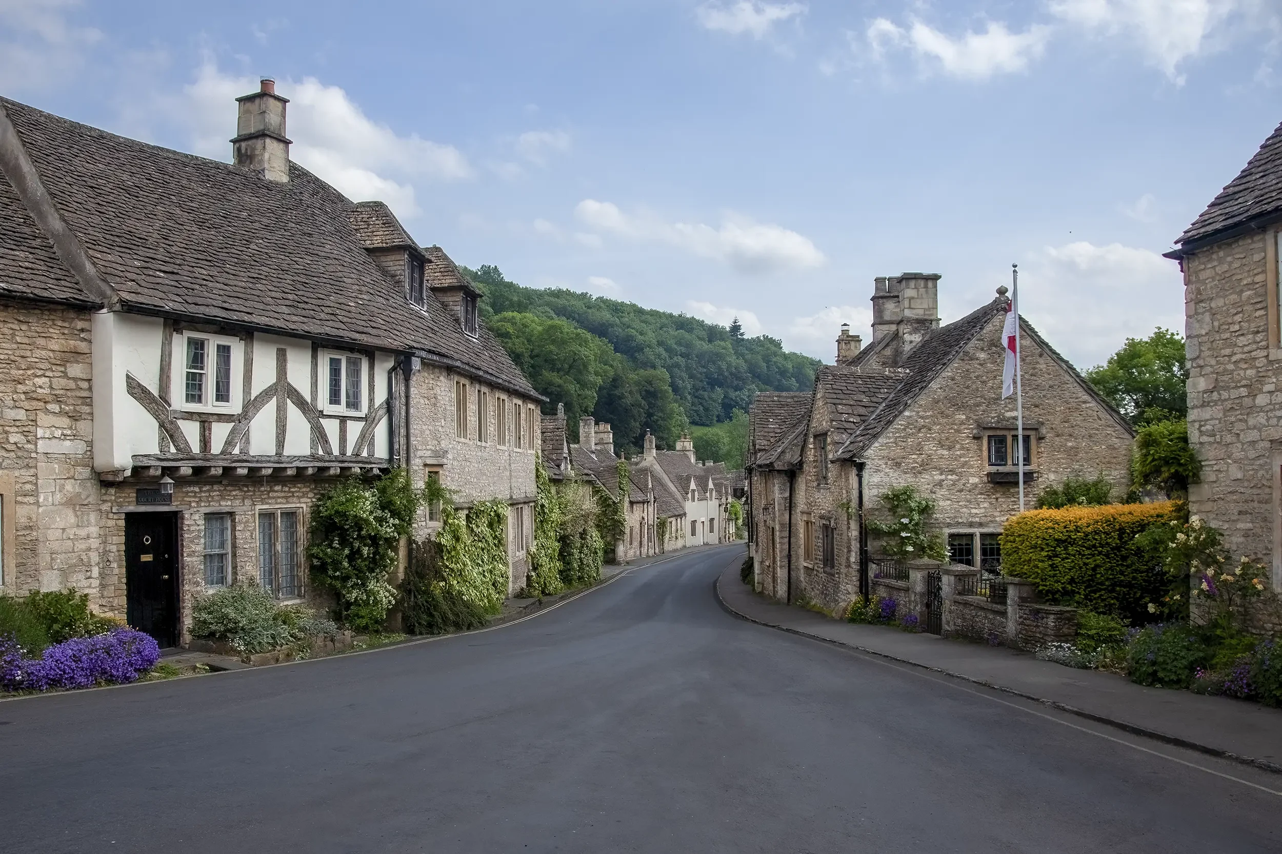 A quaint street lined with historic stone cottages with steeply pitched roofs, green foliage, and a flagpole with a white and red flag, under a partly cloudy sky in a small village setting.
