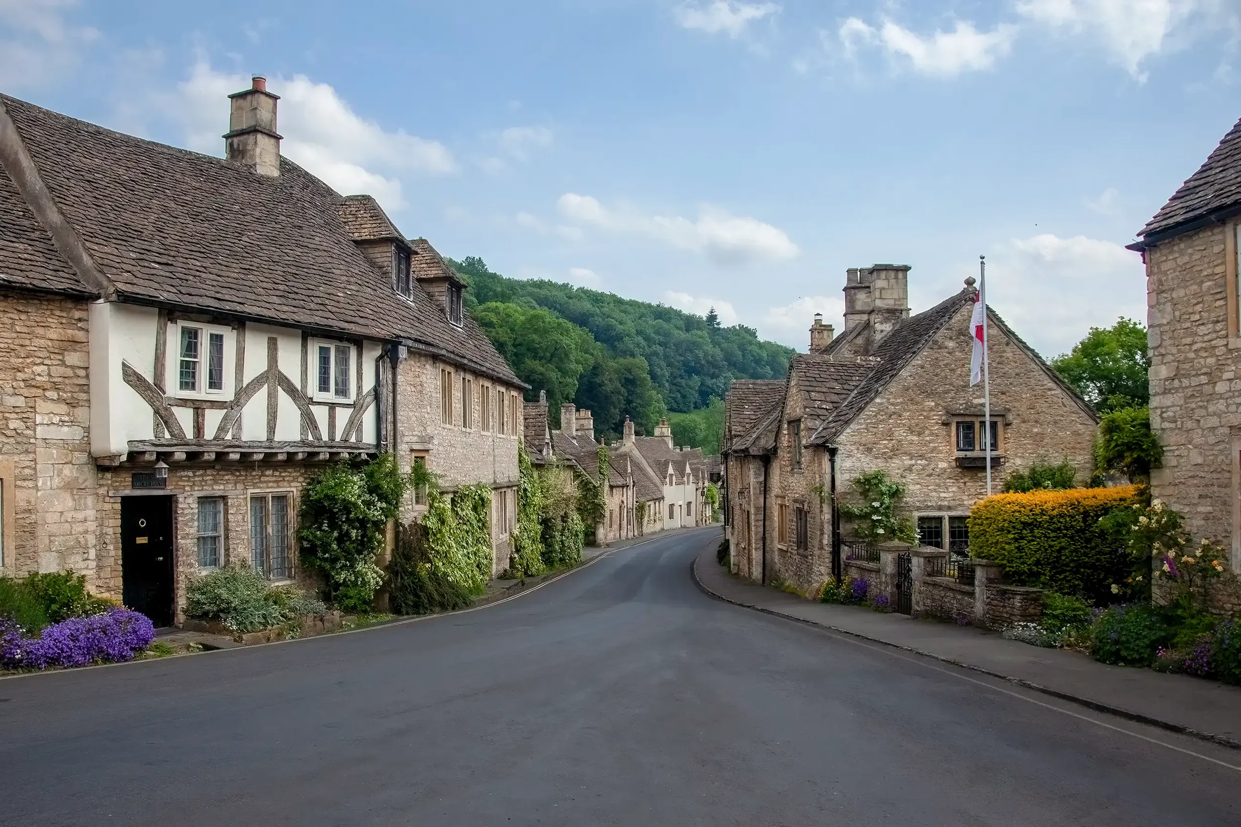 A quiet street in a small village with historic stone cottages, colorful flower beds, a flagpole with a white flag, and green hills in the background under a partly cloudy sky.