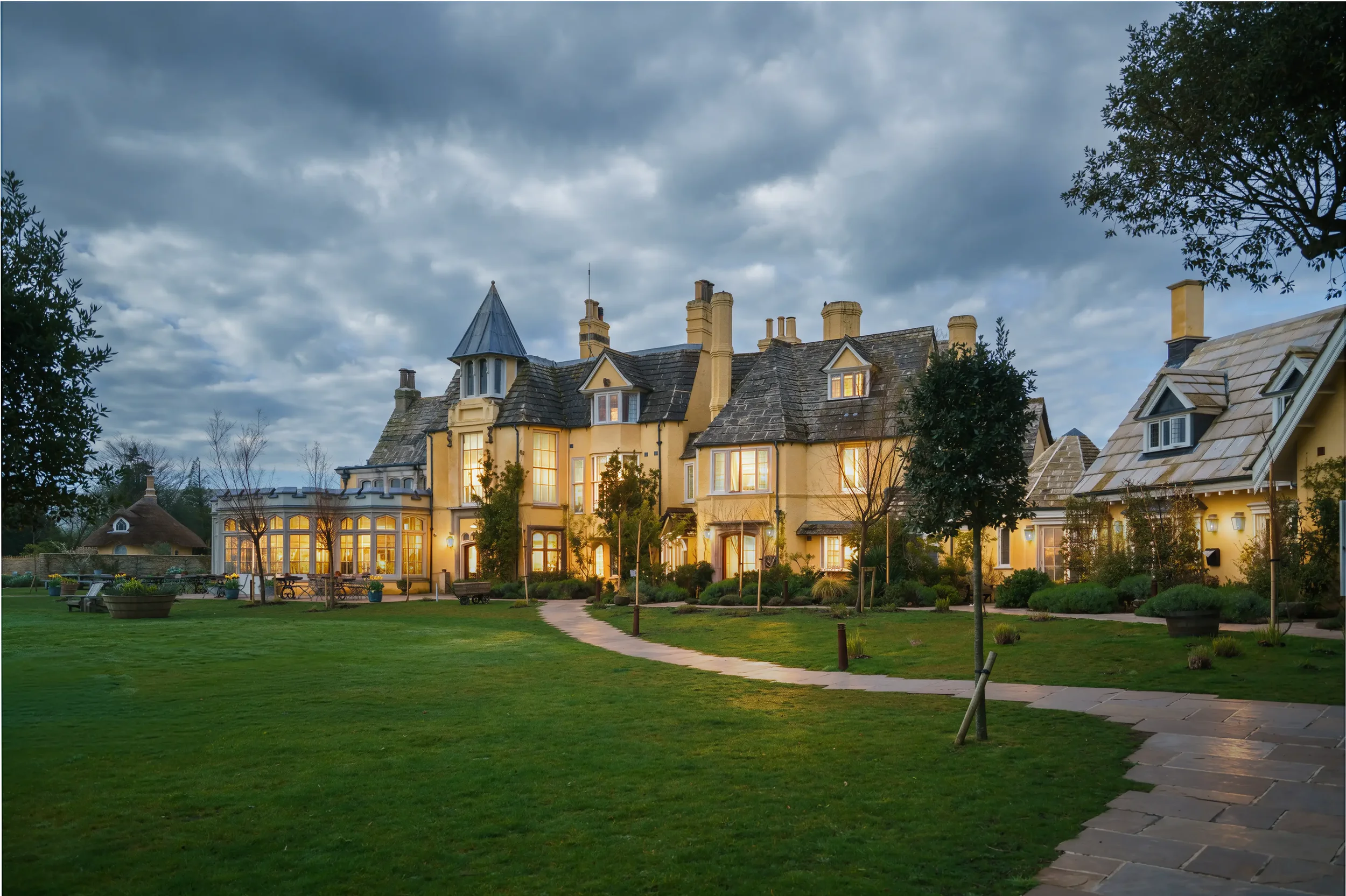 A large, elegant multi-story house with a turret, multiple chimneys, and lit windows, surrounded by a well-maintained lawn, trees, and a winding stone pathway under a cloudy evening sky.