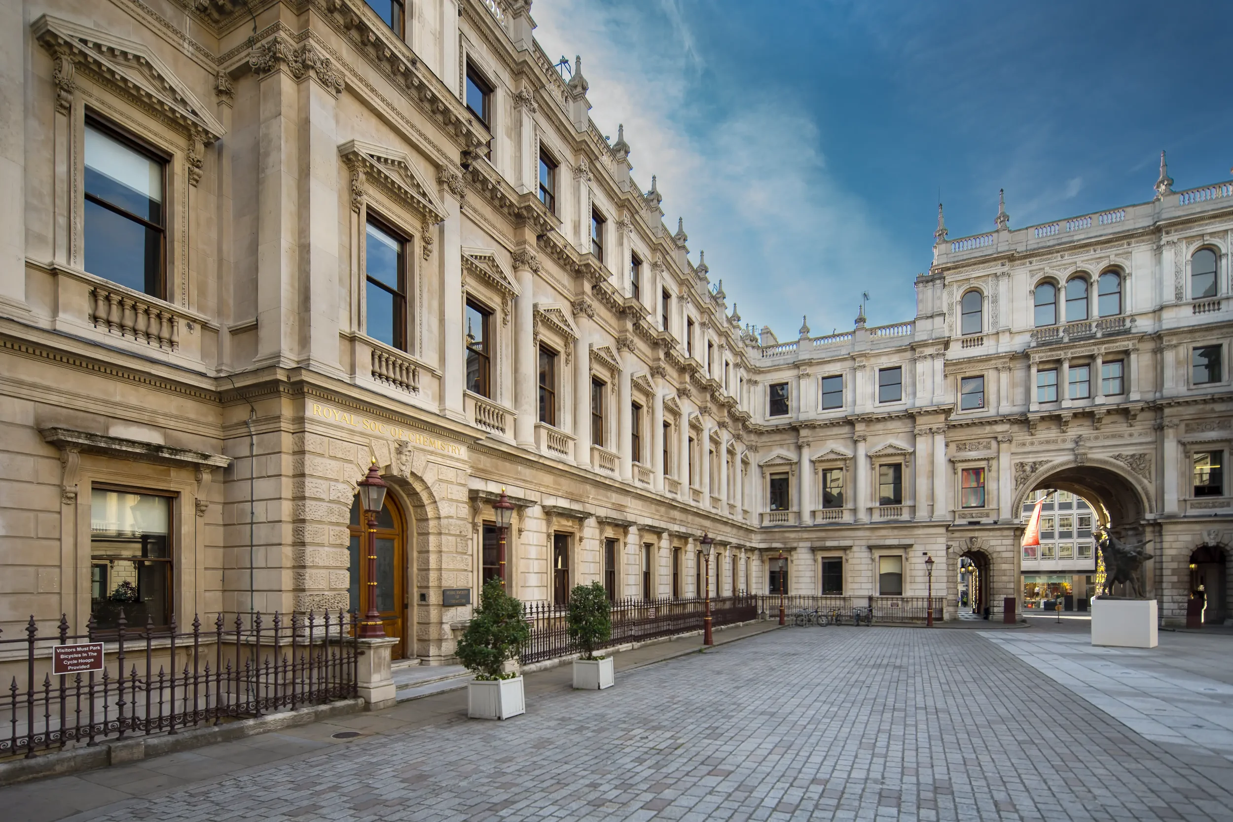 Historical building with classical architecture including detailed window trims, decorative cornices, and stone carvings, with a sky visible in the background, situated along a cobblestone street with small trees in white planters and lampposts.