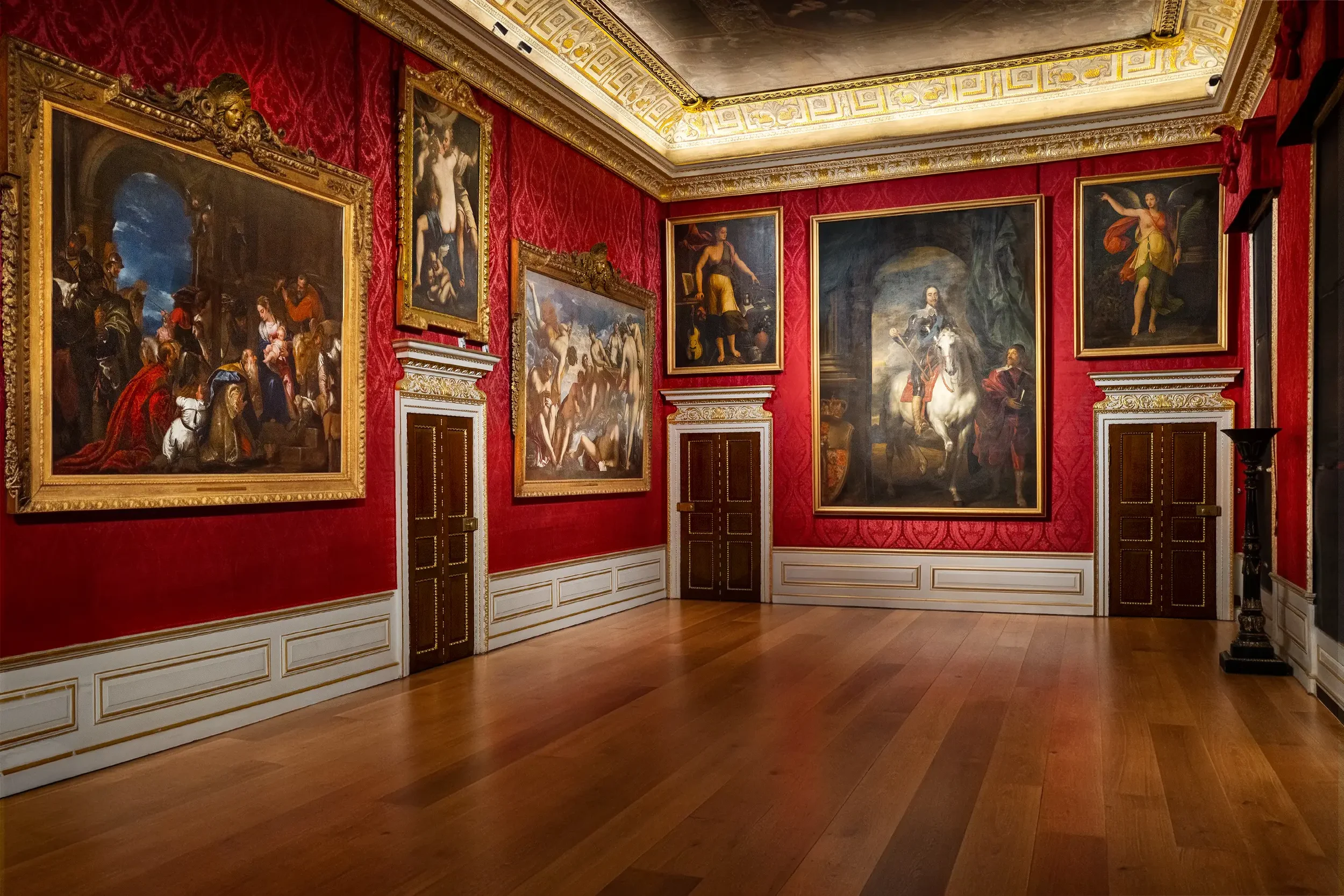 An art gallery with red patterned walls and ornate gold framing around large classical paintings, a decorative molding on the ceiling, and wooden flooring.