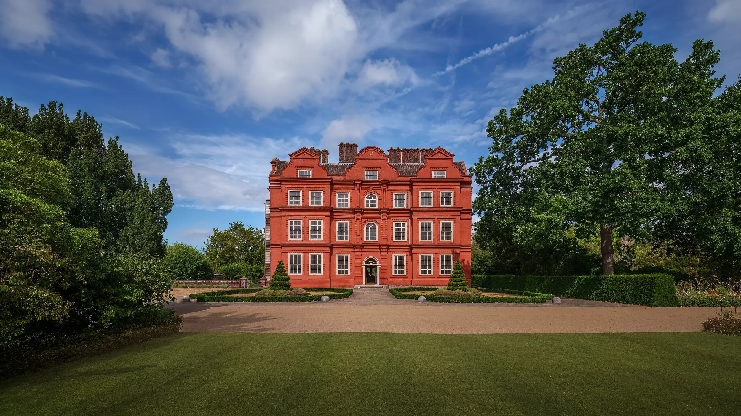 A large, red-brick building with multiple windows and ornate architectural details, surrounded by a well-manicured lawn and lush green trees, under a blue sky with scattered clouds.