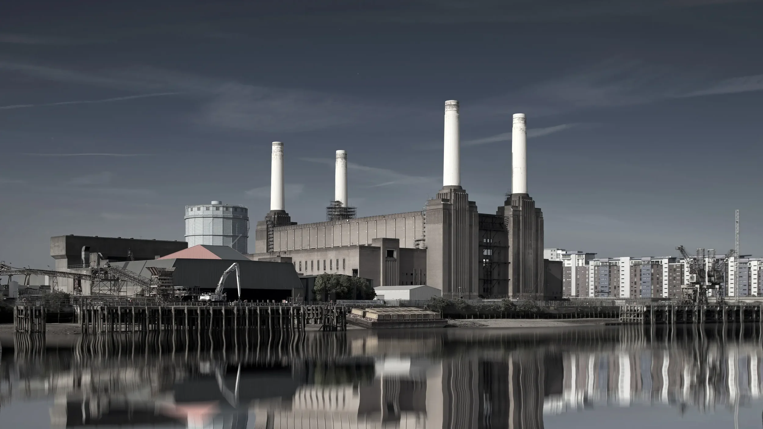 An industrial building with four tall white smokestacks, set against a dark cloudy sky, with reflection in the water below.