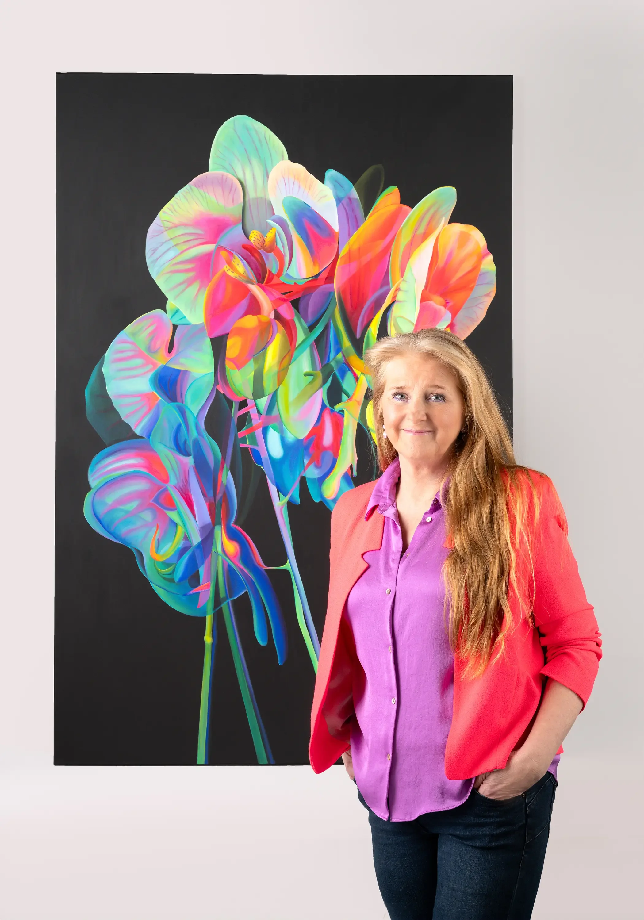 A woman with long red hair, wearing a pink blazer and purple shirt, standing in front of a large colorful painting of abstract flowers on a black background.